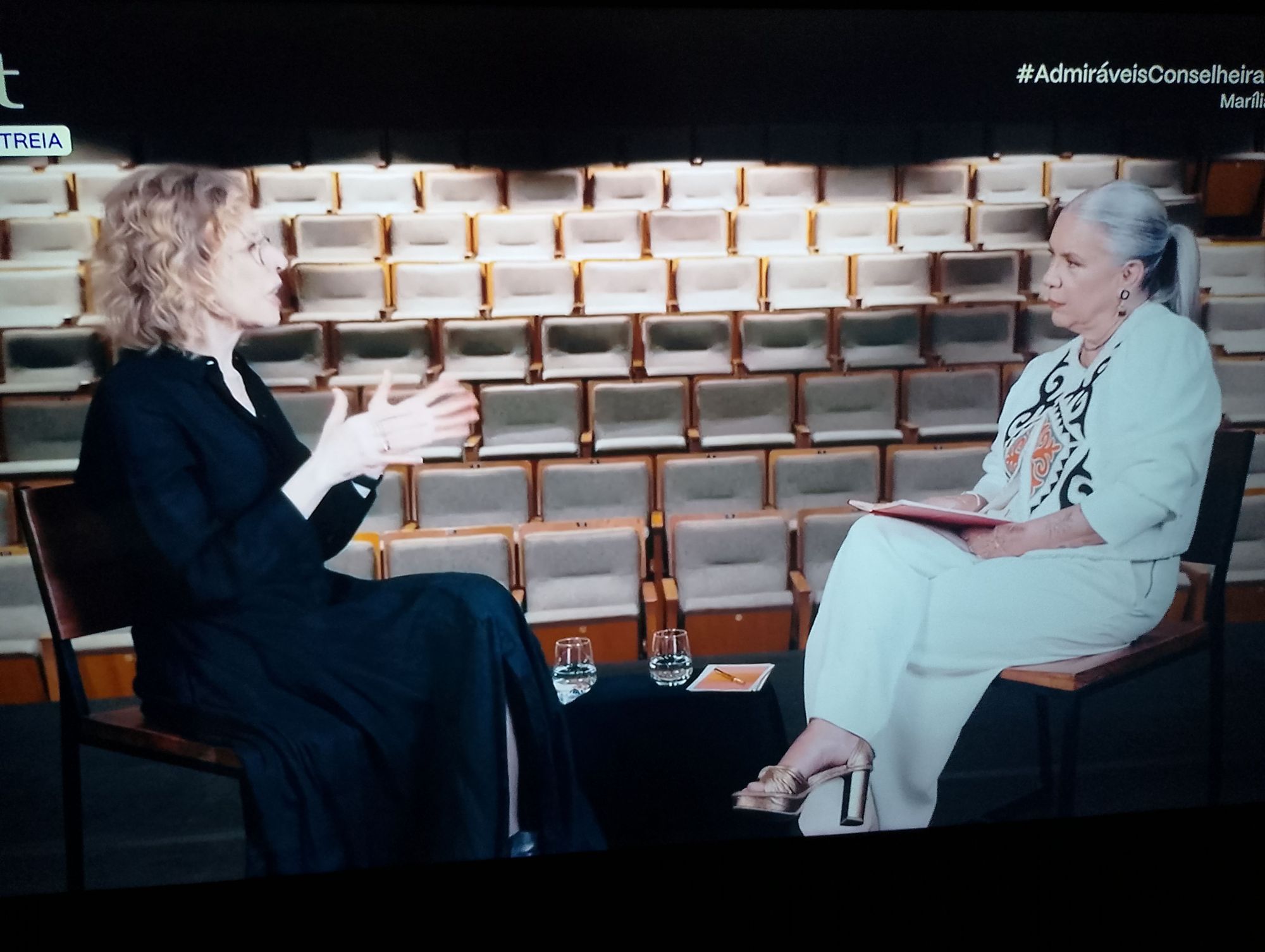 Foto da Marília Gabriela (usando roupa preta) e Astrid Fontenelle (usando roupa branca com estampa) sentadas cada uma em uma cadeira conversando.