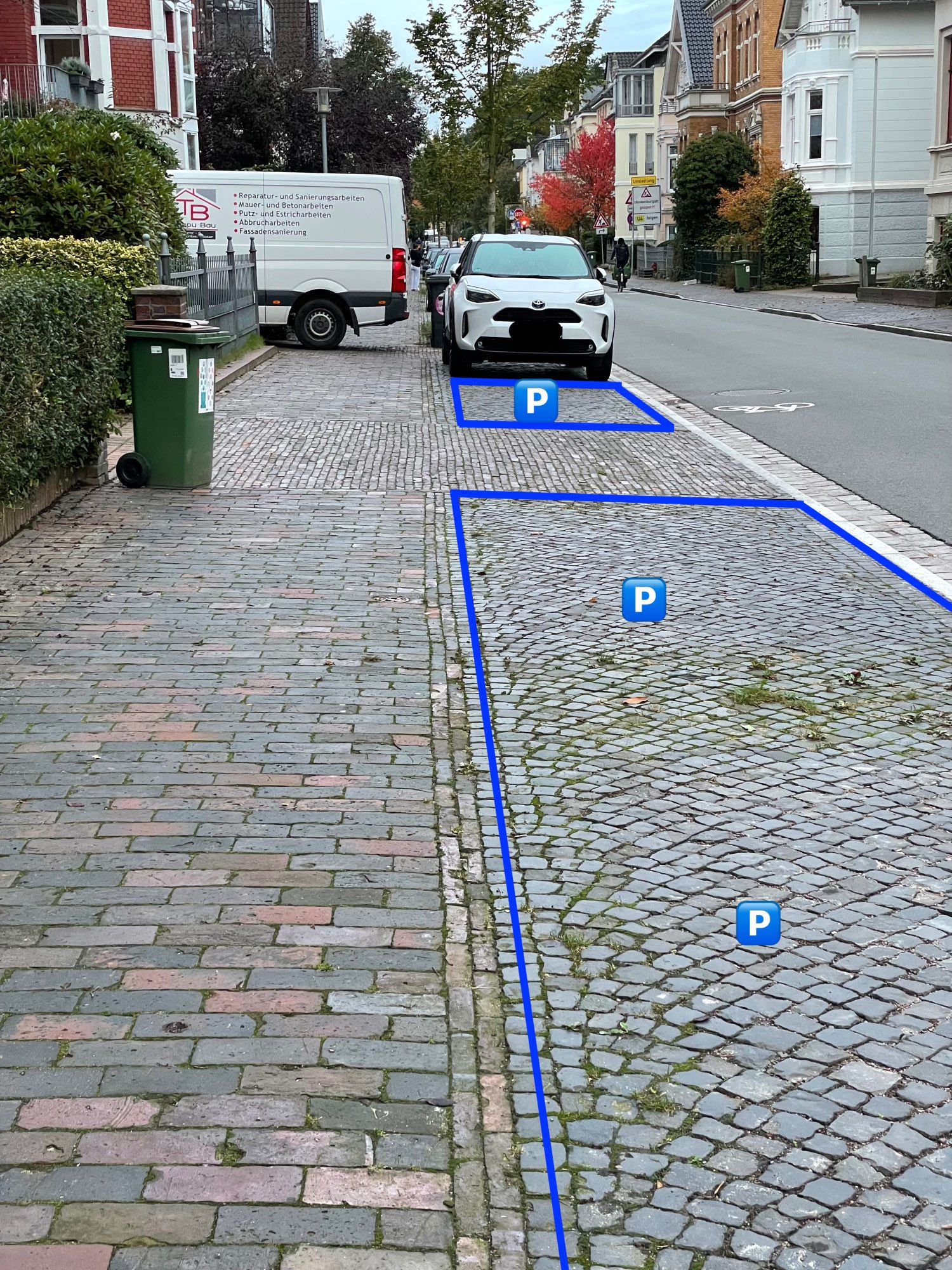 Gehweg, Parkstreifen, Fahrbahn. Im Vordergrund sind mehrere freie Parkplätze. Im Hintergrund sieht man einen Lieferwagen, der so in einer Hauseinfahrt geparkt ist, so dass sein Heck den Gehweg blockiert.