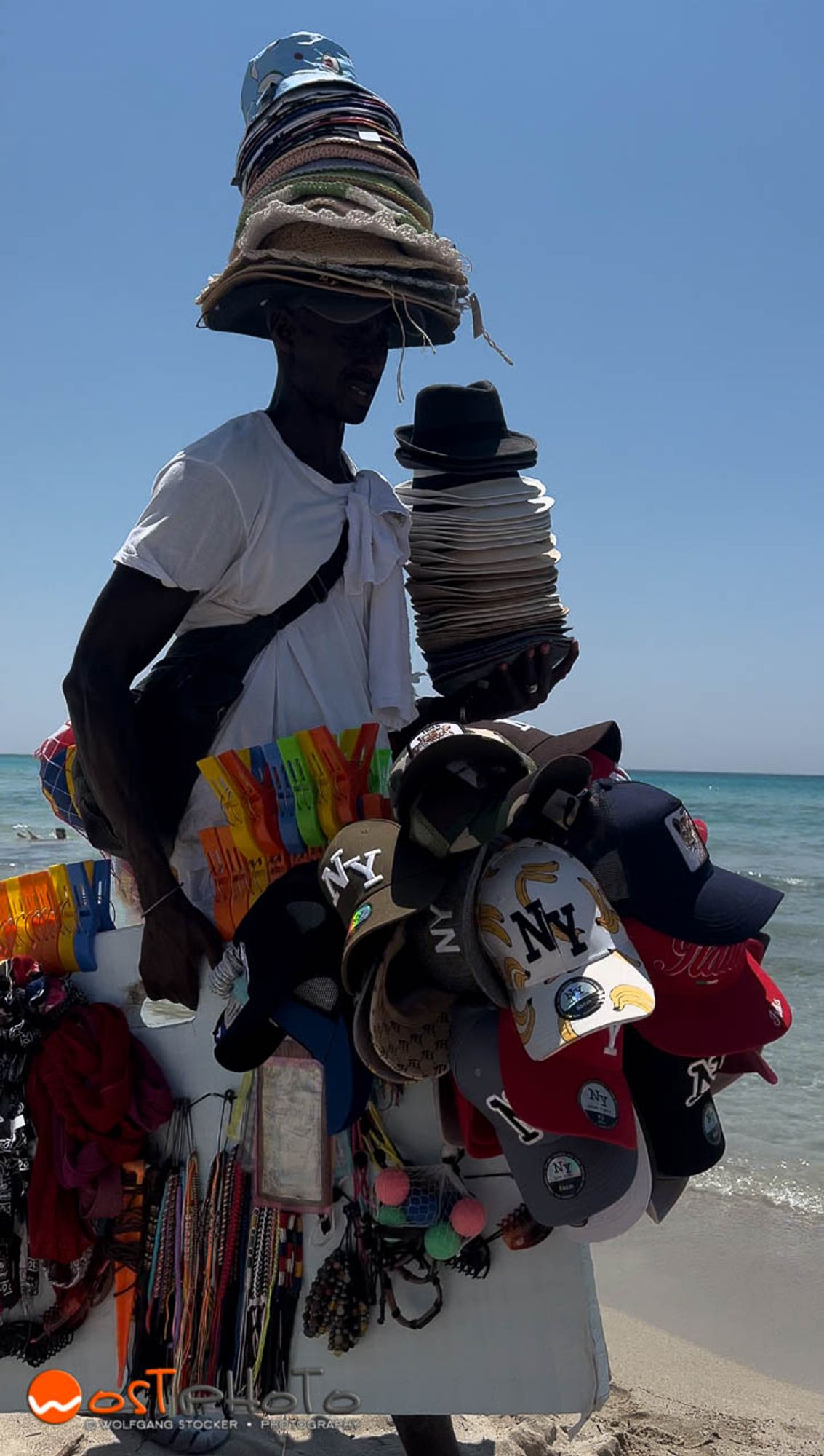 A beach vendor in Apulia in Italy, walking on the beach with multiple hats on his head and carrying all kinds of things