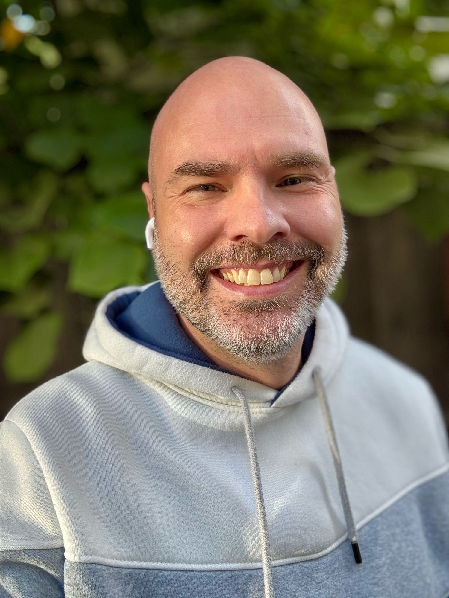 Smoothheaded smiling at the camera. He’s bald, salt and pepper beard, wearing a hoodie and there is greenery in the background.