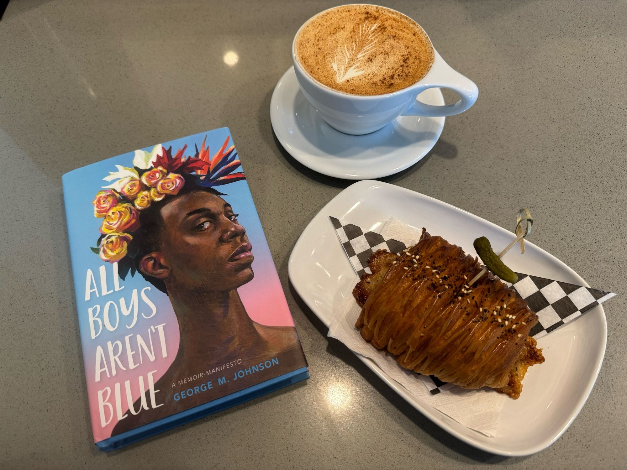 Tableau of Smoothheaded’s morning: the book, “All Boys Aren’t Blue” by George M. Johnson, a turmeric ginger chai latte, and a ham & Gruyère croissant.