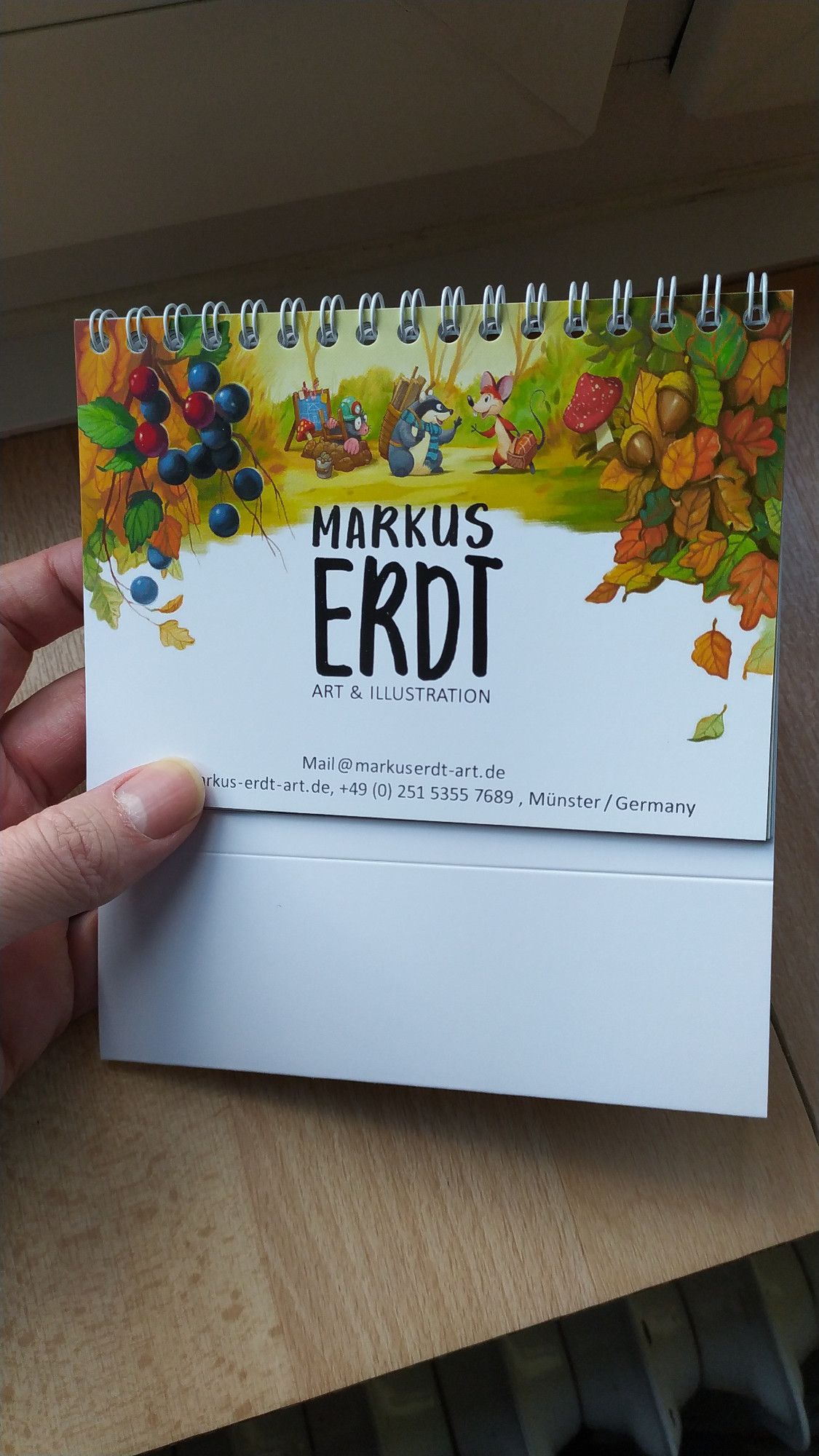 A small desktop calendar as a handout for clients. The cover shows a fantasy autumn scene on the top half with cute critters in the middle, nuts, berries and leaves around the corners. In the middle, the name of the designer: Markus Erdt. Contact at the bottom.