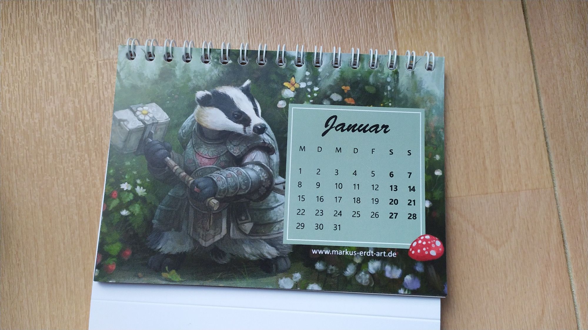 A page of a desktop calendar. The month, days and numbers are presented on a mint green square on the right, while the background is filled with a fantasy illustration of a badger in armor, wielding a hammer decorated with flowers and strawberries. He stands in a dark green, lush forest scene. Art by Markus Erdt.