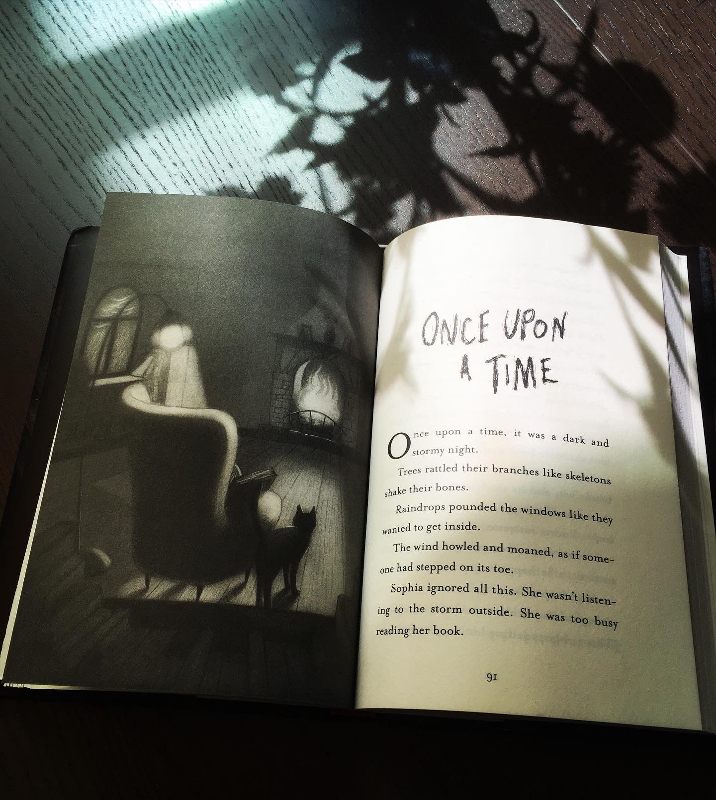 Book open to a full-page illustration by Carolina Godina of a black cat next to an empty arm chair in front of a fireplace, opposite the first page of the story Once Upon a Time. The book is on a wooden table, with shadows from a vase of plants cast over it 