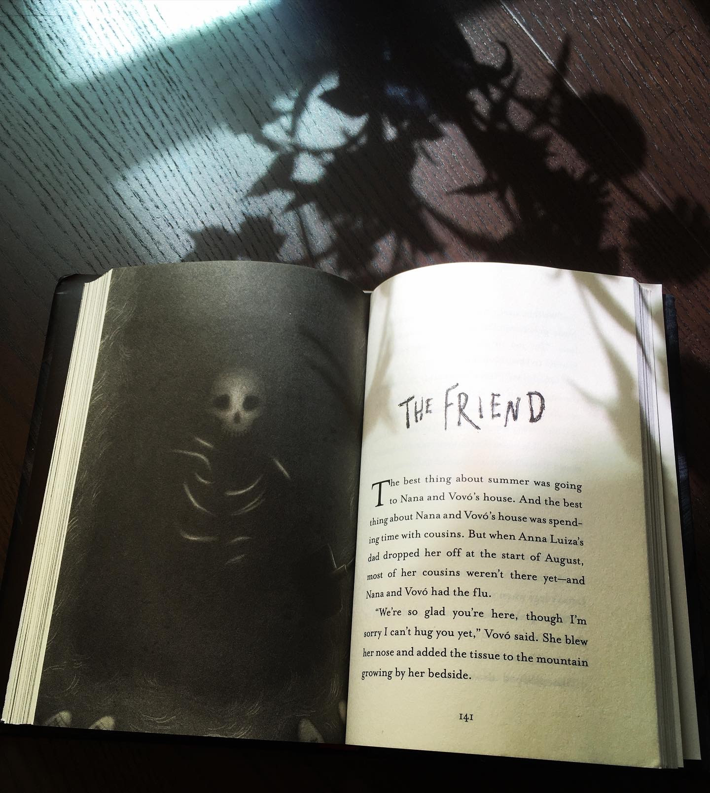 Book open to a full-page illustration by Carolina Godina of a skeleton in the ground, with a kid's sneakers visible at the edge of the dug-up grave, opposite the first page of the story The Friend. The book is on a wooden table, with shadows from a vase of plants cast over it 