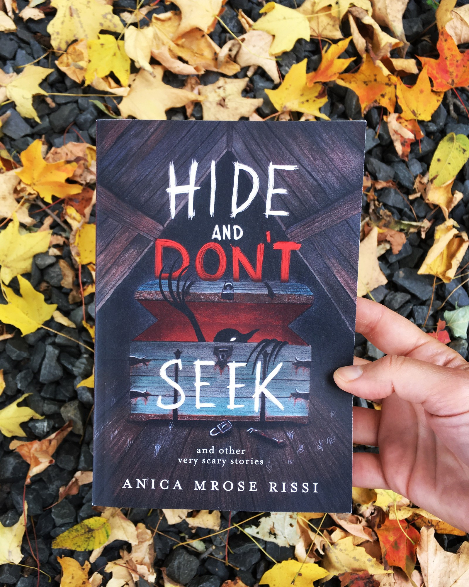 Paperback of HIDE AND DON'T SEEK: AND OTHER VERY SCARY STORIES by Anica Mrose Rissi, held up over a background of rocks and yellow/orange leaves. Cover illustration by Carolina Godina shows a creature coming out of a trunk in an attic. Spooky!