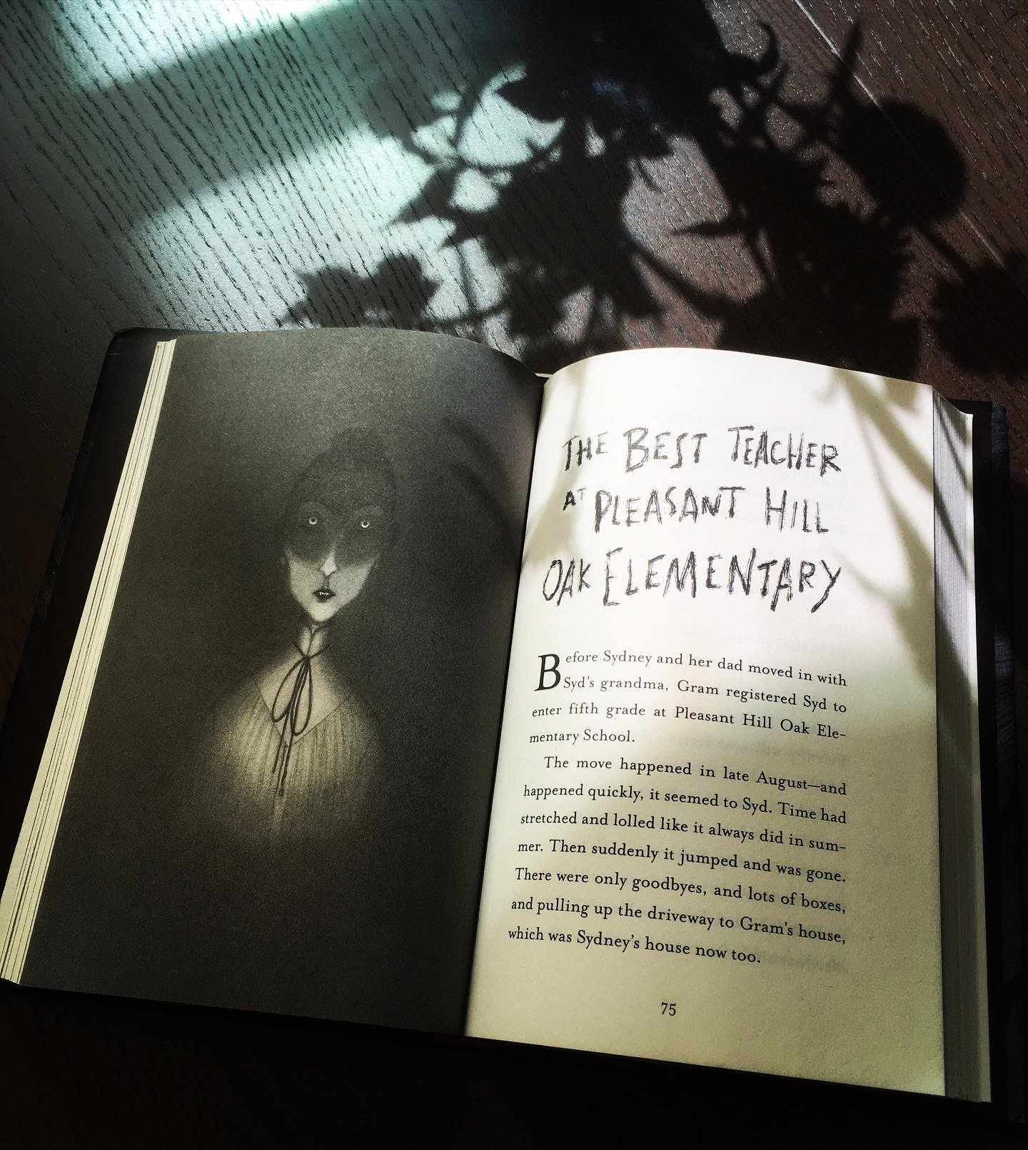 Book open to a full-page illustration by Carolina Godina of a spooky teacher in shadow, opposite the first page of the story The Best Teacher at Pleasant Hill Oak Elementary. The book is on a wooden table, with shadows from a vase of plants cast over it 