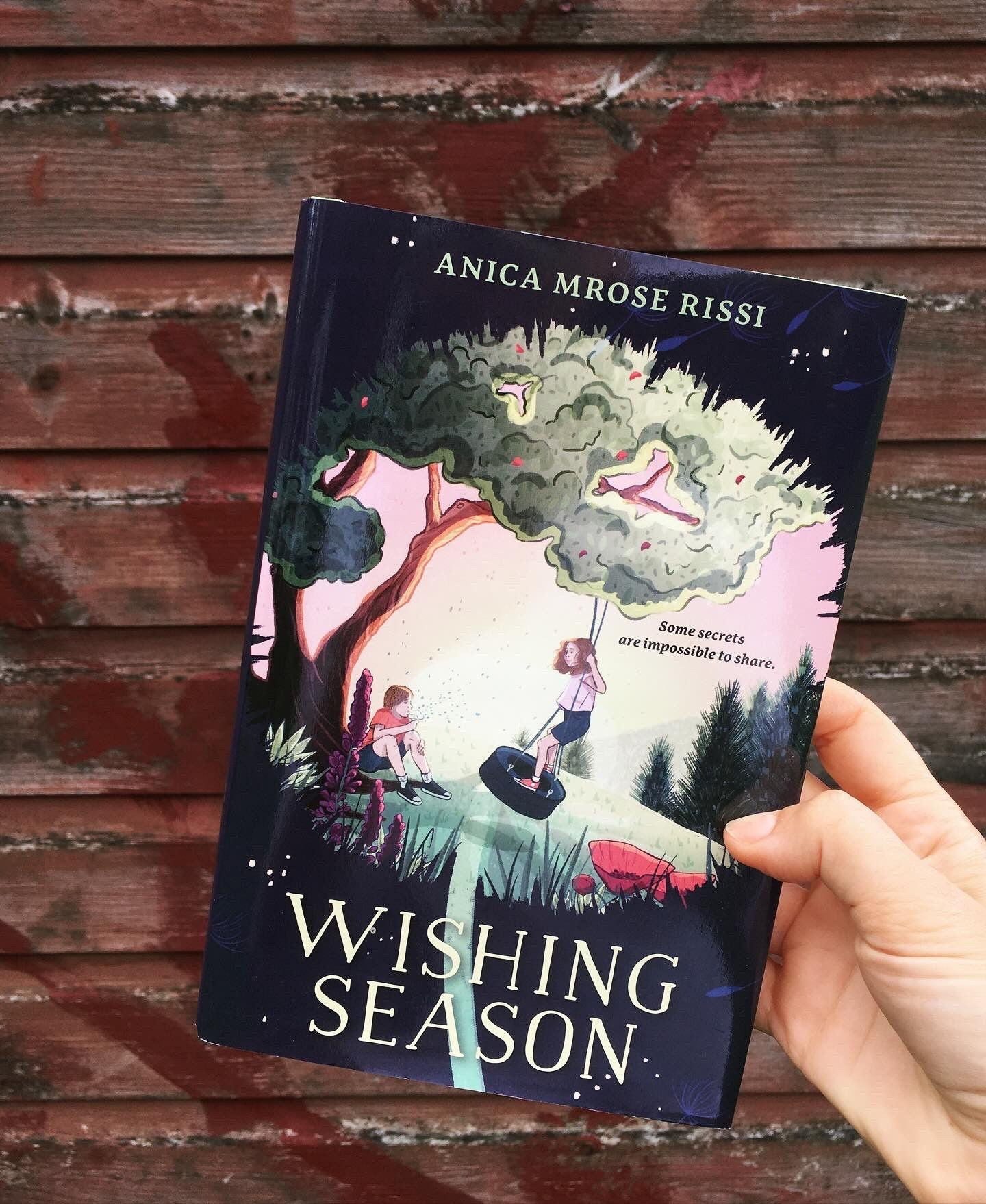 Hardcover of WISHING SEASON by Anica Mrose Rissi held up in front of a faded red-painted wooden wall