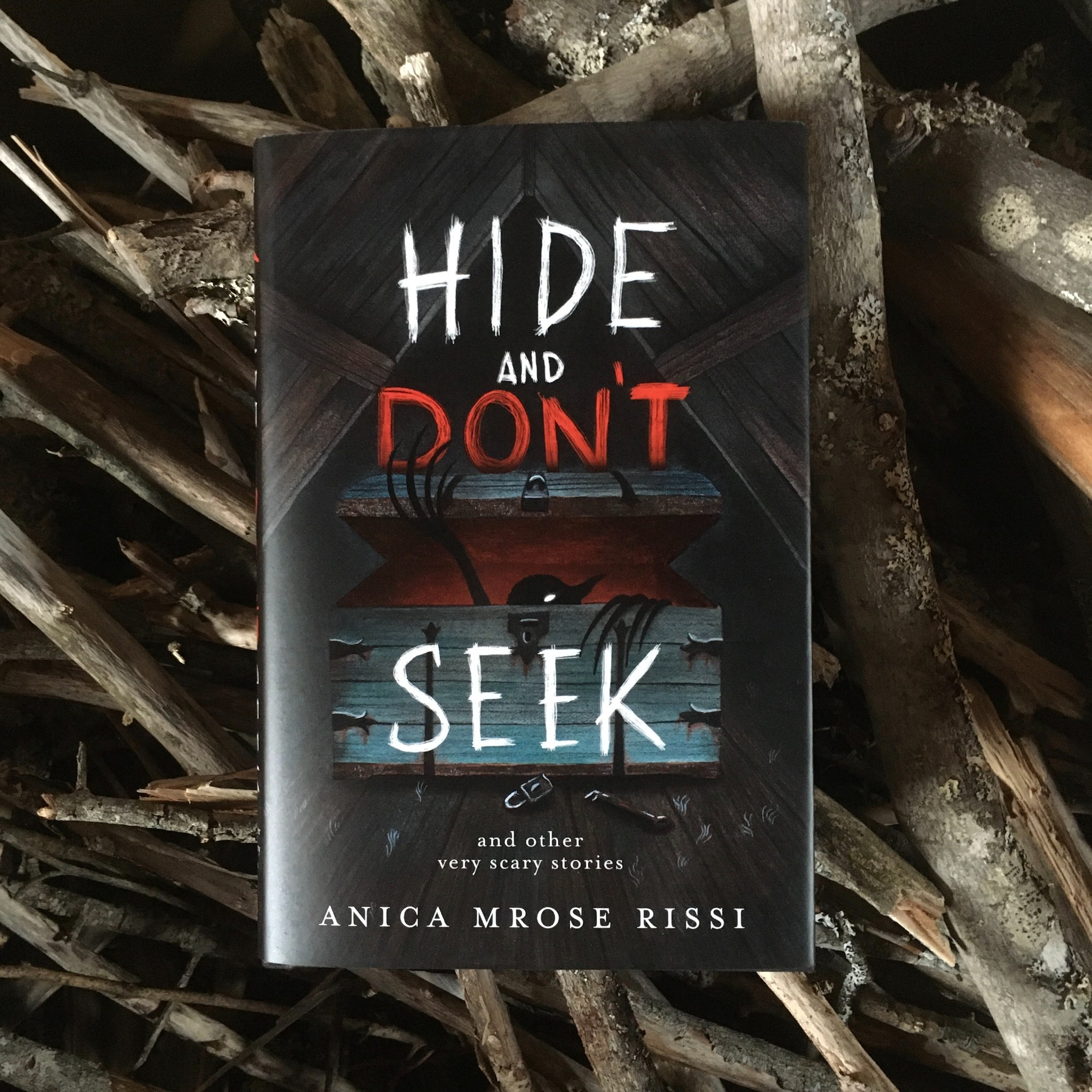 Hardcover of HIDE AND DON'T SEEK: AND OTHER VERY SCARY STORIES by Anica Mrose Rissi, on top of a pile of sticks. Cover illustration by Carolina Godina shows a creature emerging from a trunk in an attic.