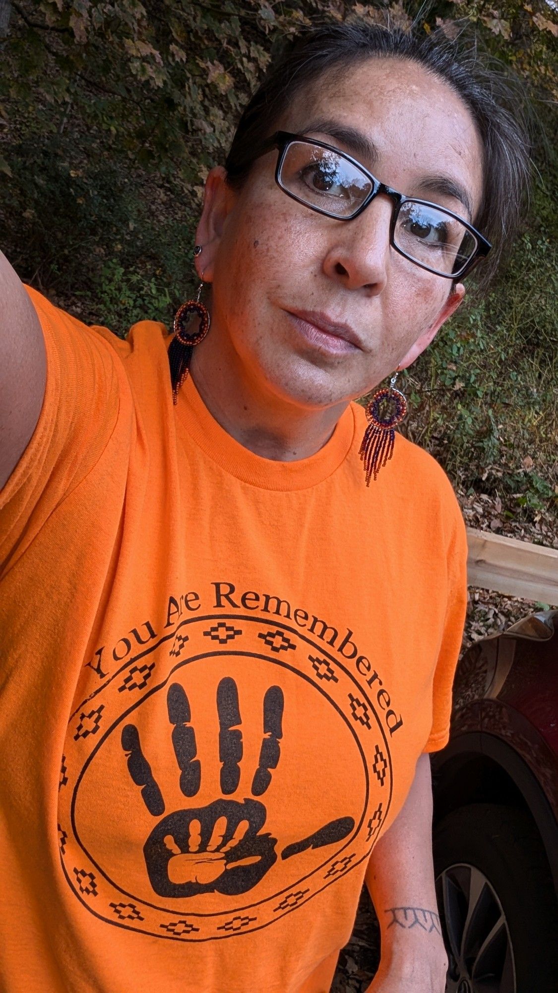 Woman with glasses wears an orange T-shirt that sayd You Are Remembered. She also has orange and blue earrings that match the shirt