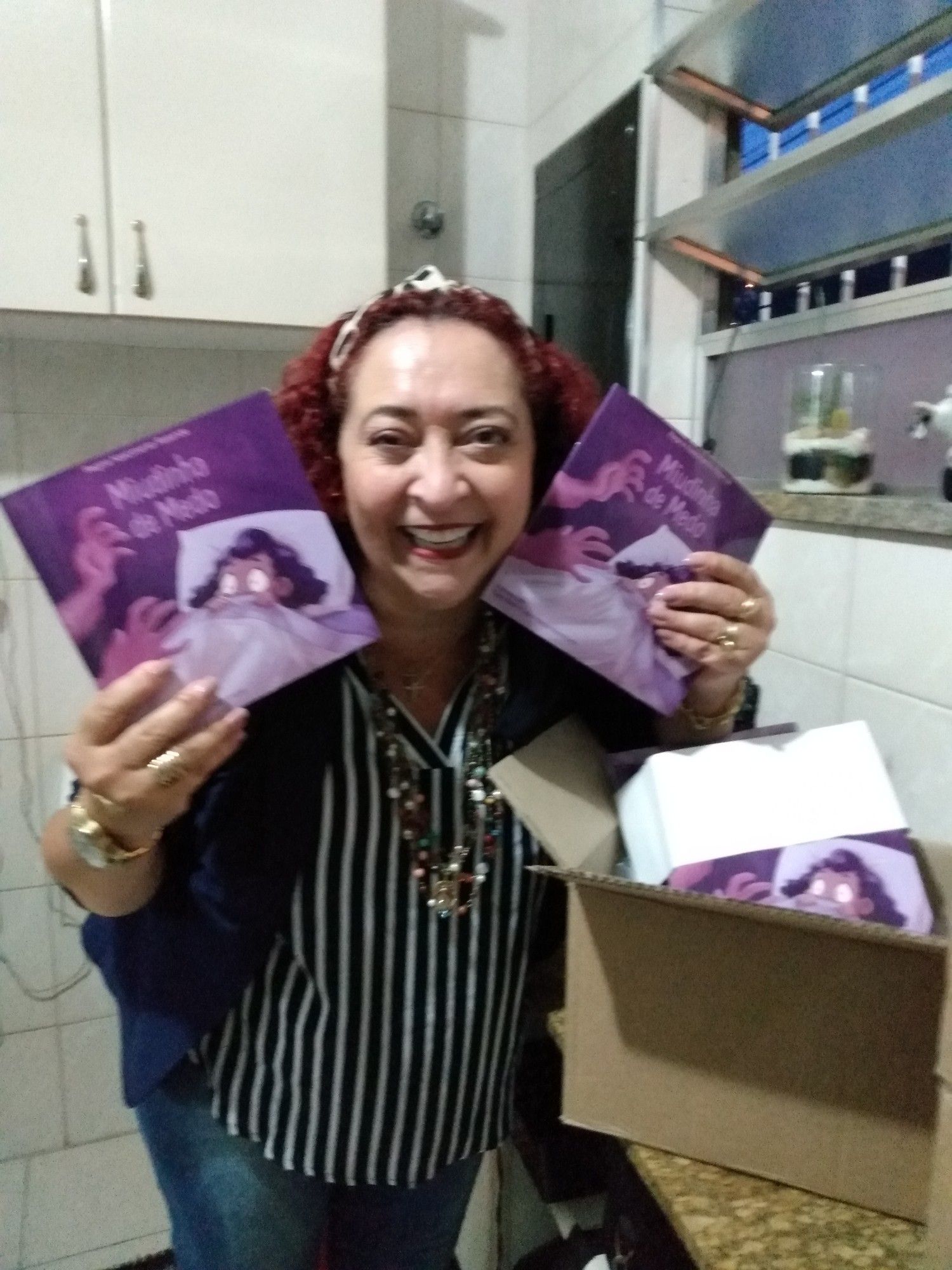 Foto de minha mãe sorrindo com uma caixa dos livros lançados. Na capa o título "Miudinha de Medo", em cor roxa com uma menina na cama com medo de mãos que se aproximam dela no escuro.