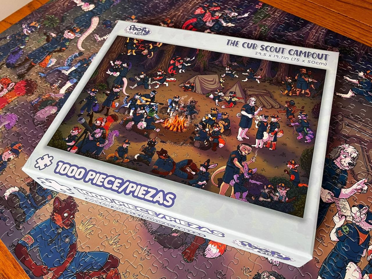 Image shows a jigsaw puzzle box, sitting on top of the finished puzzle. The illustration on the puzzle is a large Cub Scout furry troop out in the woods for a campout.