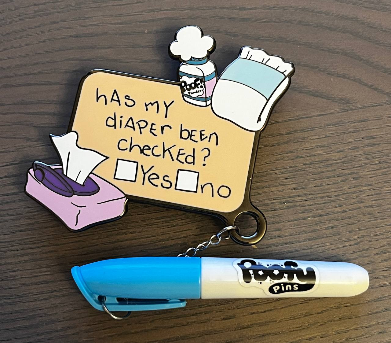 Photo is of an enamel pin that says "has my diaper been checked" with two boxes for yes and no. The pin has a miniature blue marker attached to it. The images on the pin are a fresh diaper, an open package of wipes with one pulled and a bottle of baby powder that has a cloud poofing out. On the pen is the logo "Poofy Pins".