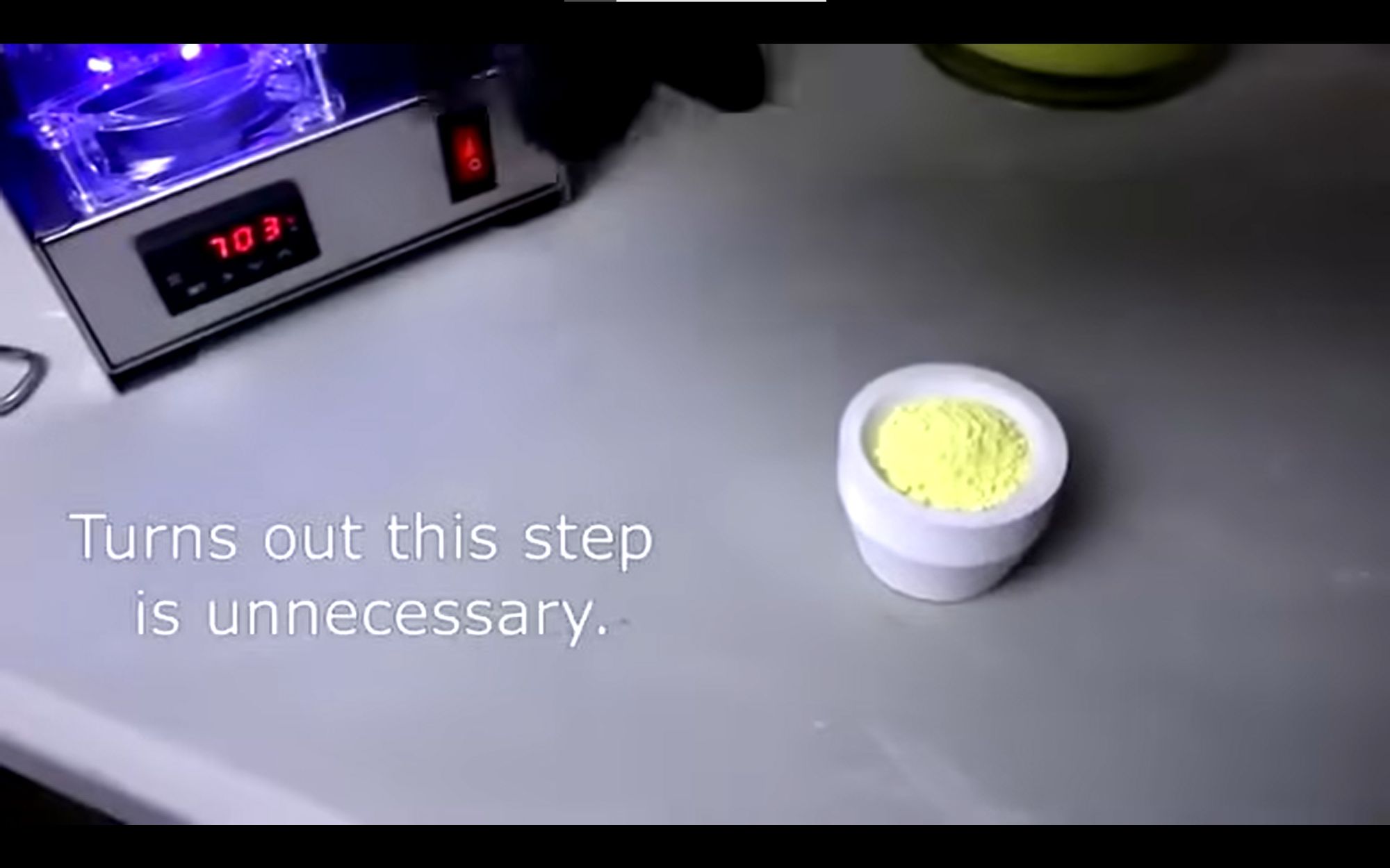 screenshot of the infamous codyslab uranium refinement video showing a small crucible filled with uranium oxide, a yellow powder, on a metal table in front of equipment with a purple ultraviolet glow. text says "Turns out this step is unnecessary."