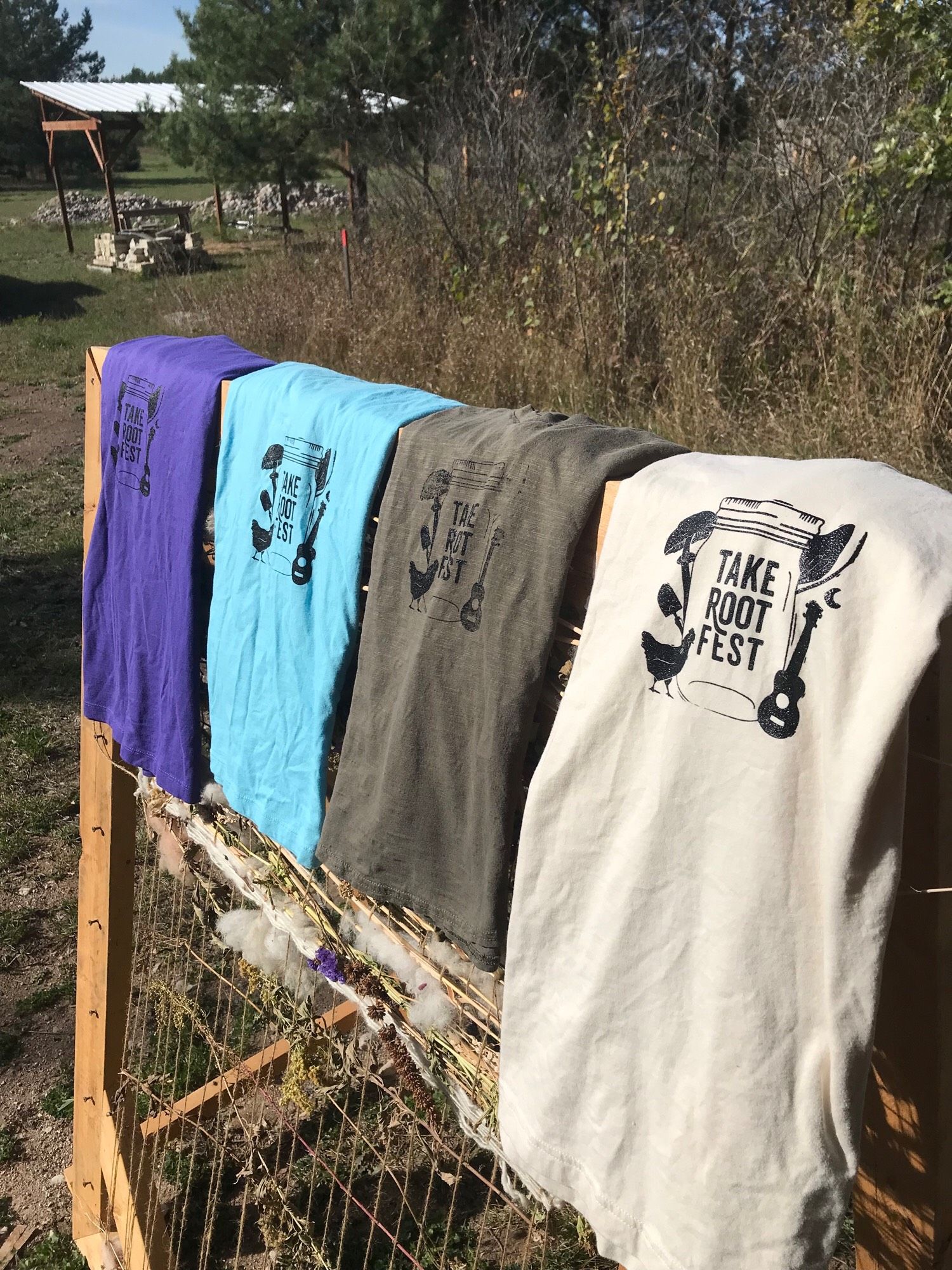 Four different-coloured shirts hanging on a “natural weaving loom” outside; they each have a freshly-screen-printed “Take Root Fest” logo on them, in black. (Did I ever mentioned we also run a festival?)