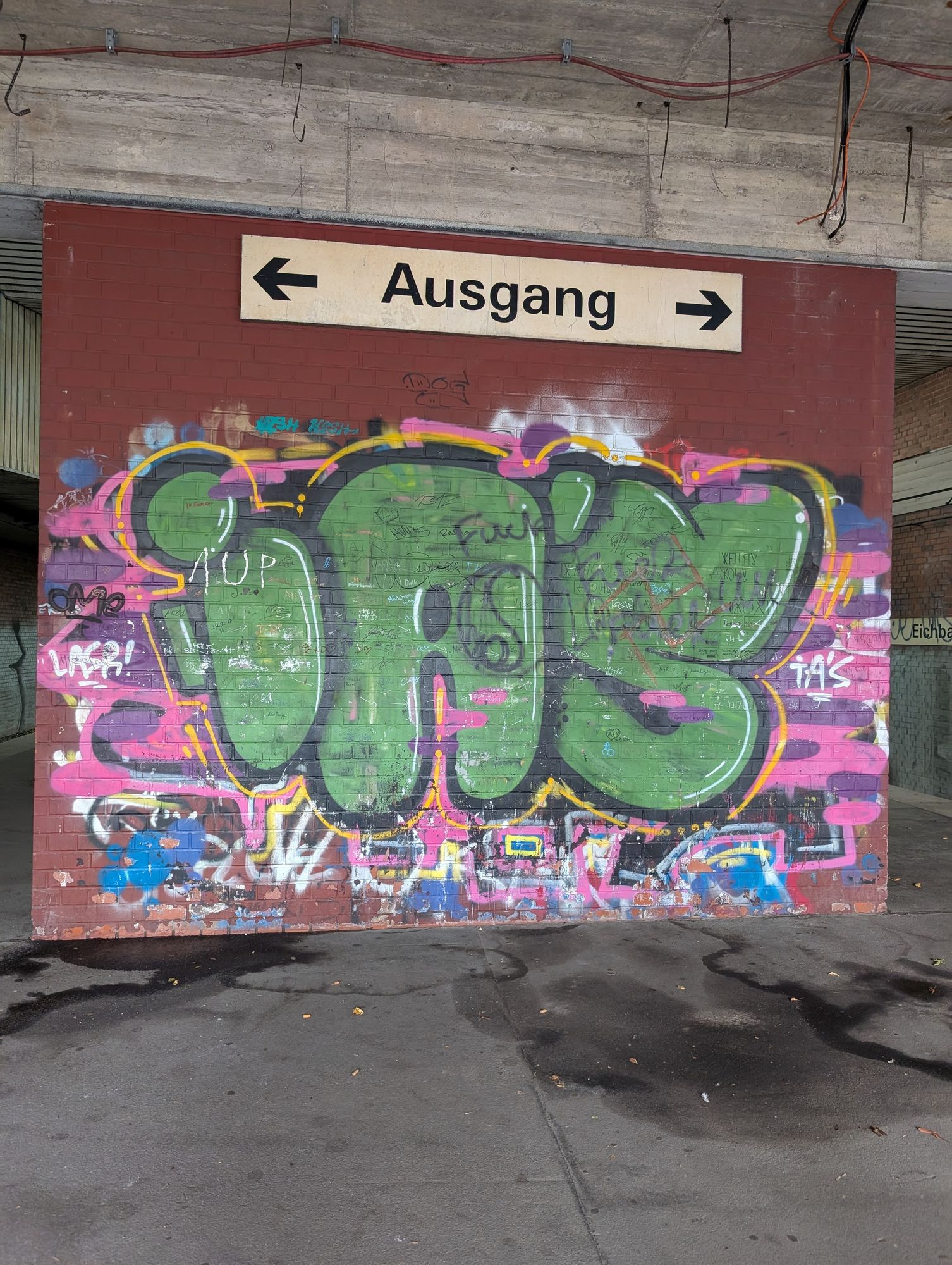 Kunterbunt besprayte und beschriebene Wand.
Darüber ein Schild auf dem steht Ausgang mit Pfeilen nach rechts und links 