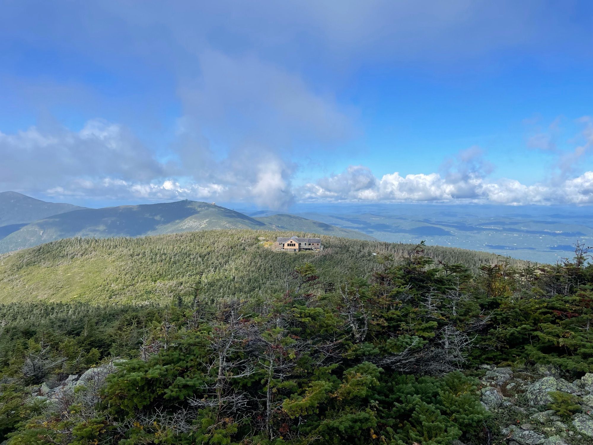 Photograph of the Appalachian Mountain Club Greenleaf Hut with Cannon Mountain and the Kinsmans in the middle distance and the upper Connecticut River valley visible. There are some clouds in the sky but the sun is shining on the hut.