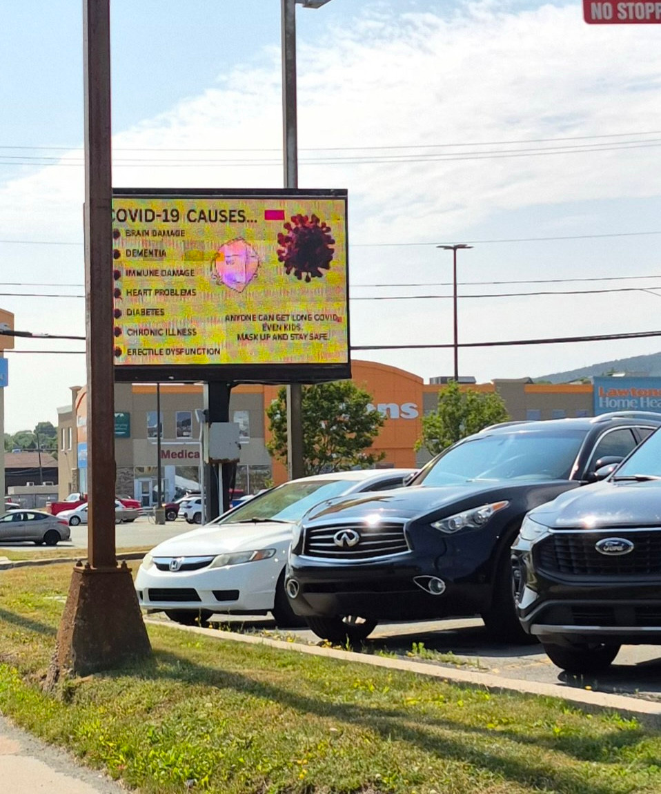 There is a large green digital billboard display with the words COVID-19 CAUSES...
BRAIN DAMAGE
DEMENTIA
IMMUNE DAMAGE
HEART PROBLEMS
DIABETES
CHRONIC ILLNESS
N95
ERECTILE DYSFUNCTION
ANYONE CAN GET LONG COVID, EVEN KIDS.
MASK UP AND STAY SAFE. On it. There is also a graphic of a purplish virus particle, and a white n95 mask. There are also 3 vehicles in the picture, two are dark blue suvs and the other is a white car.  There are buildings in the background and a patch of grass in front of the parking lot where the vehicles are. It is a sunny day.