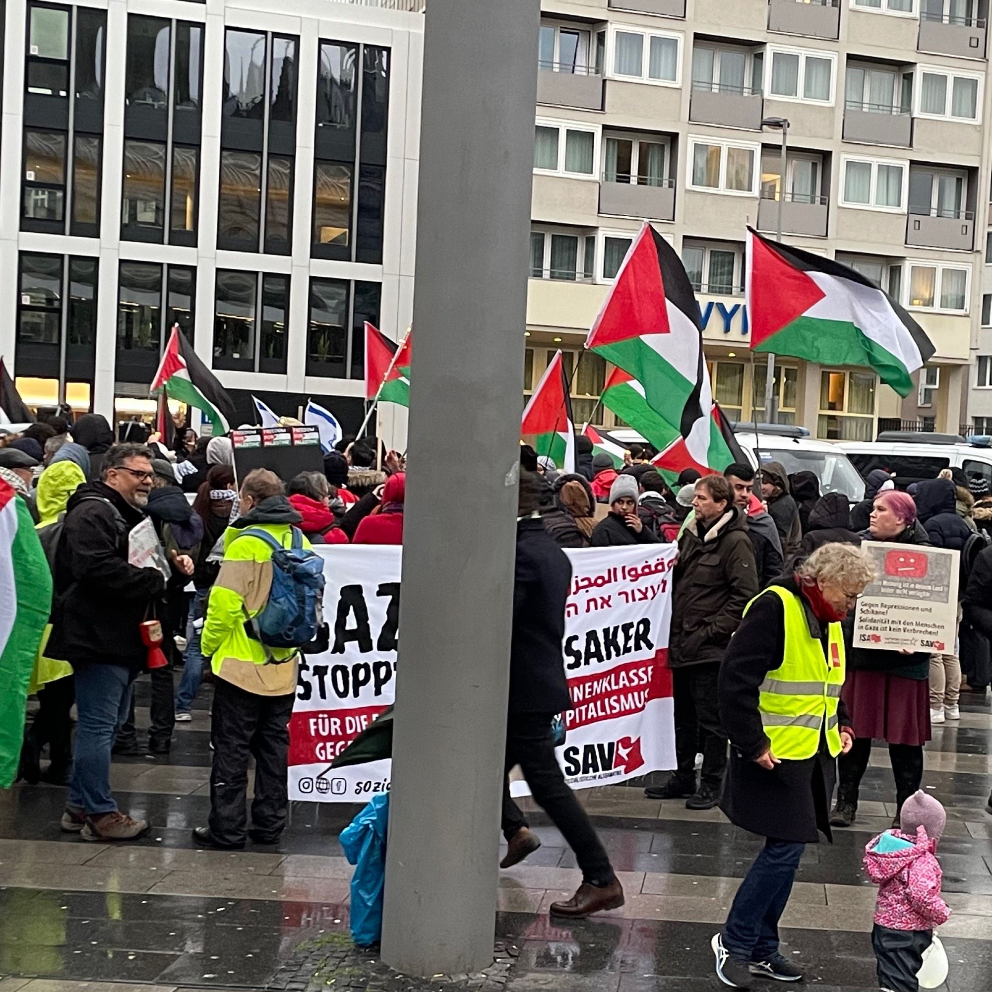 Ältere weiße Männer halten auf einer Demo das am 22.11. von der Polizei bei der Demo an der Uni verbotene Transparent mit der Aufschrift „Gaza Stopp das Massaker“. Sie umgeben von nationalistischen Palästinensischen Symbolen der hinter ihnen stehenden Menschen. So voll sieht es nicht aus.