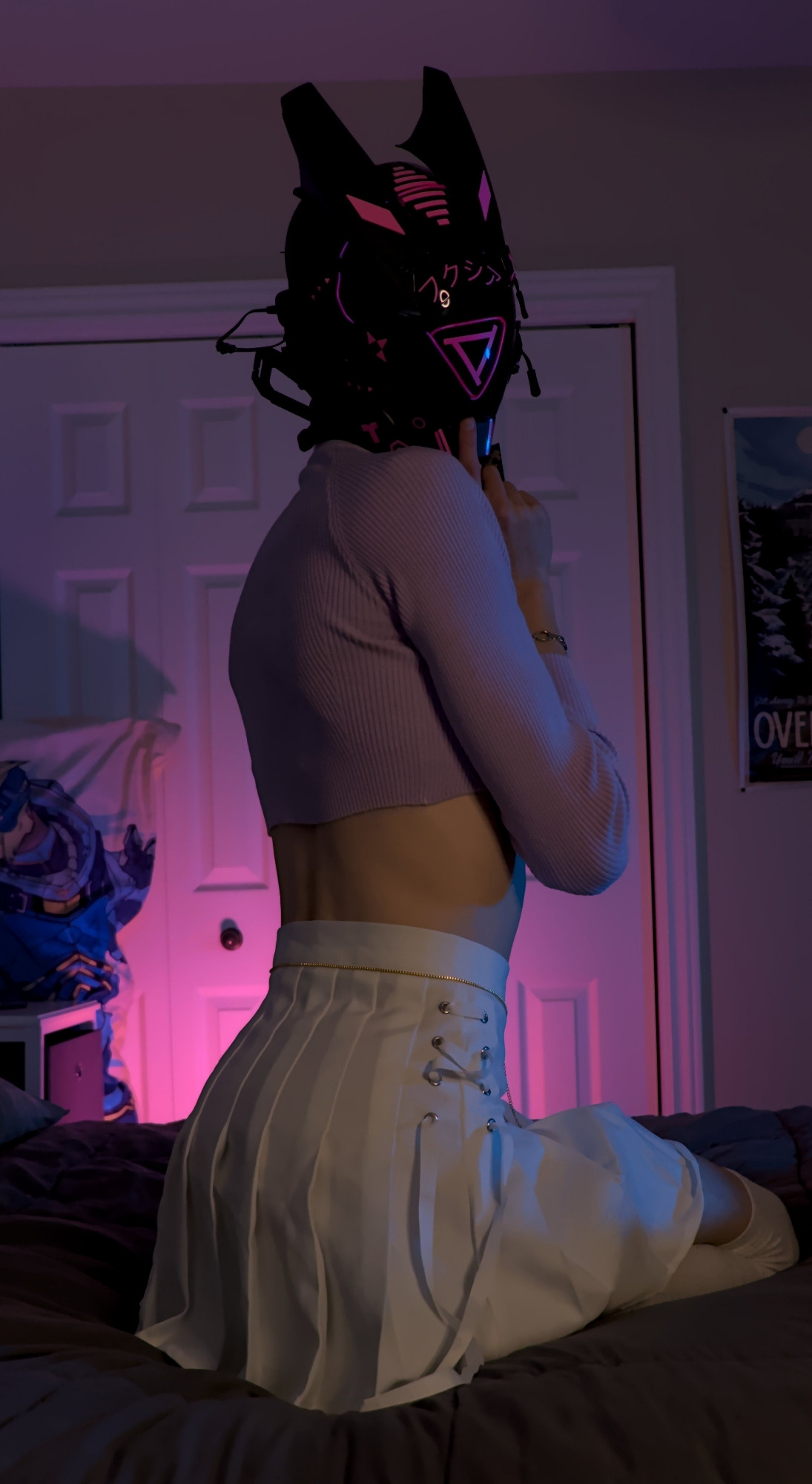 The robot Fuchsia on the bed, on his knees as his body faces away from the camera but in a side way. He's looking at the camera and wears a skirt and a crop top.