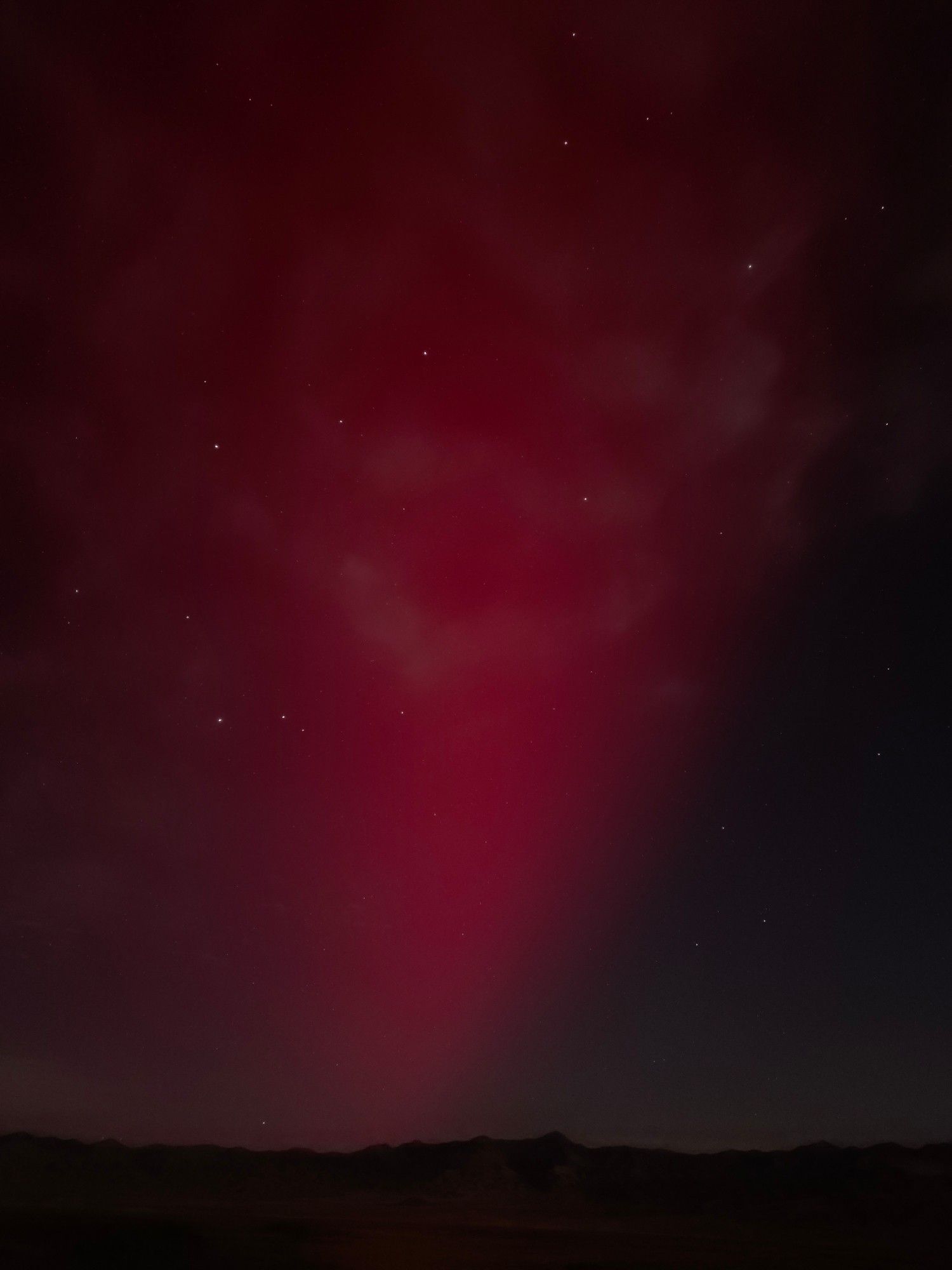 A deep red shaft of light seemingly rises from the line of black mountains, the leading edge causing a well defined separation from the pale red aurora and moonlit night sky. Stars fill the night sky behind the ghostly white clouds.