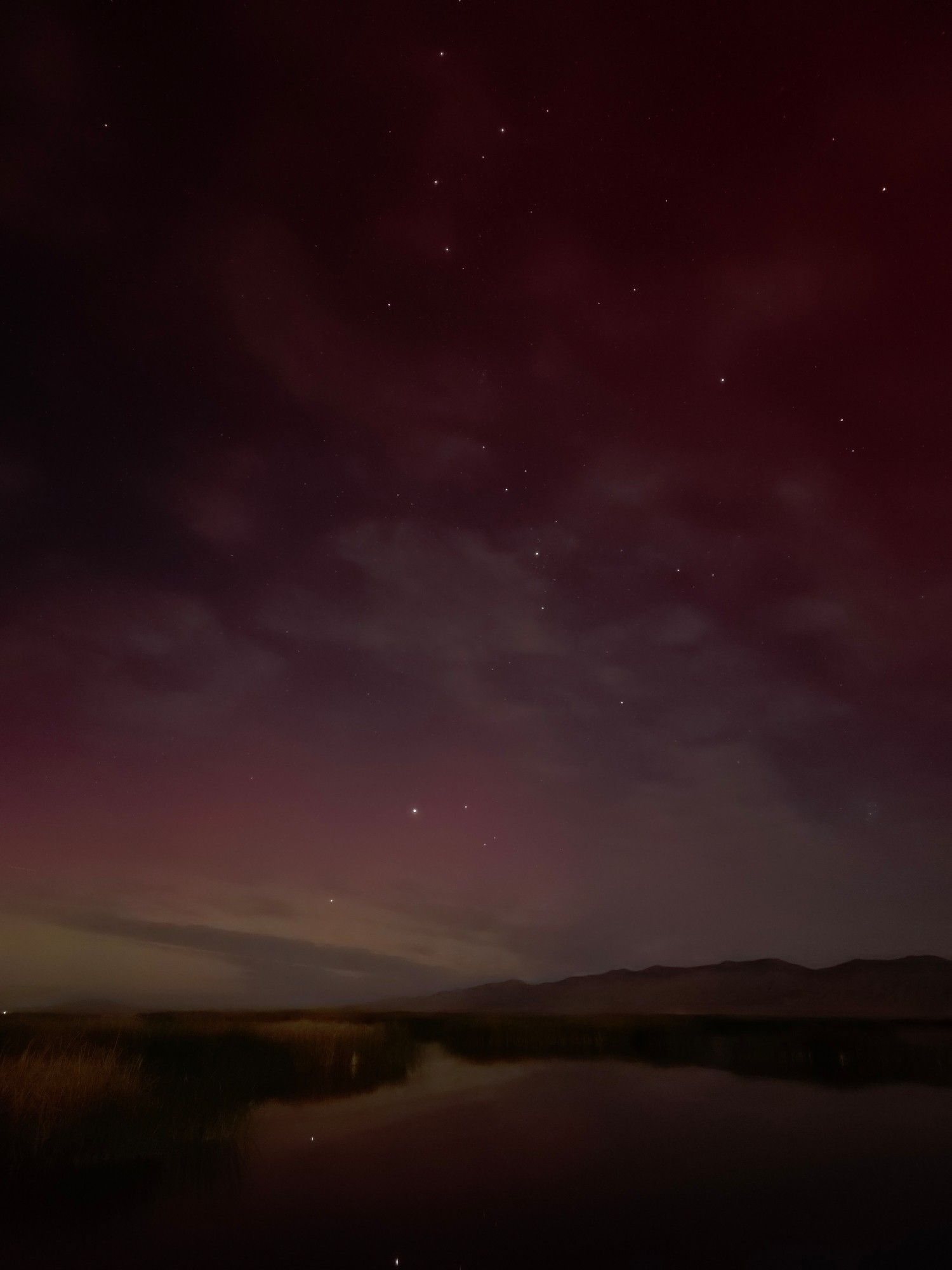 A thick line of brights stars, from the distinctive W shaped Cassiopeia down to the dark moonlit mountains below, are surrounded by a pale pink and green aurora. The brightest stars and the faint color is dimly reflected in the still lake that fills the foreground.