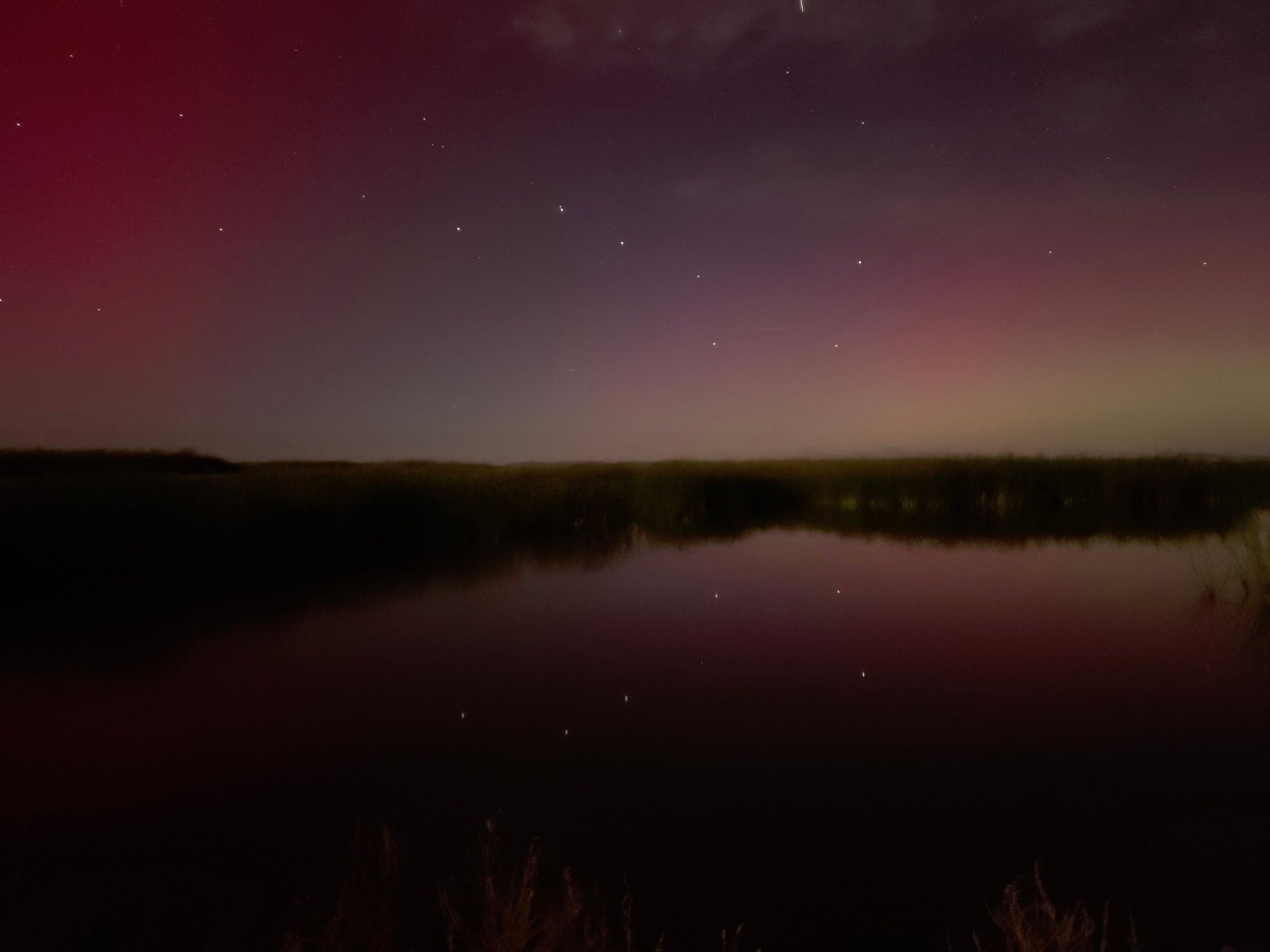 A pale pink and yellow aurora on the right fades to a dark red on the left, with the distinctive ladle shaped constellation Big Dipper centered in the image. Below it, a small lake is so calm the surface is a mirror, casting a dim reflection of the stars and colors above it.