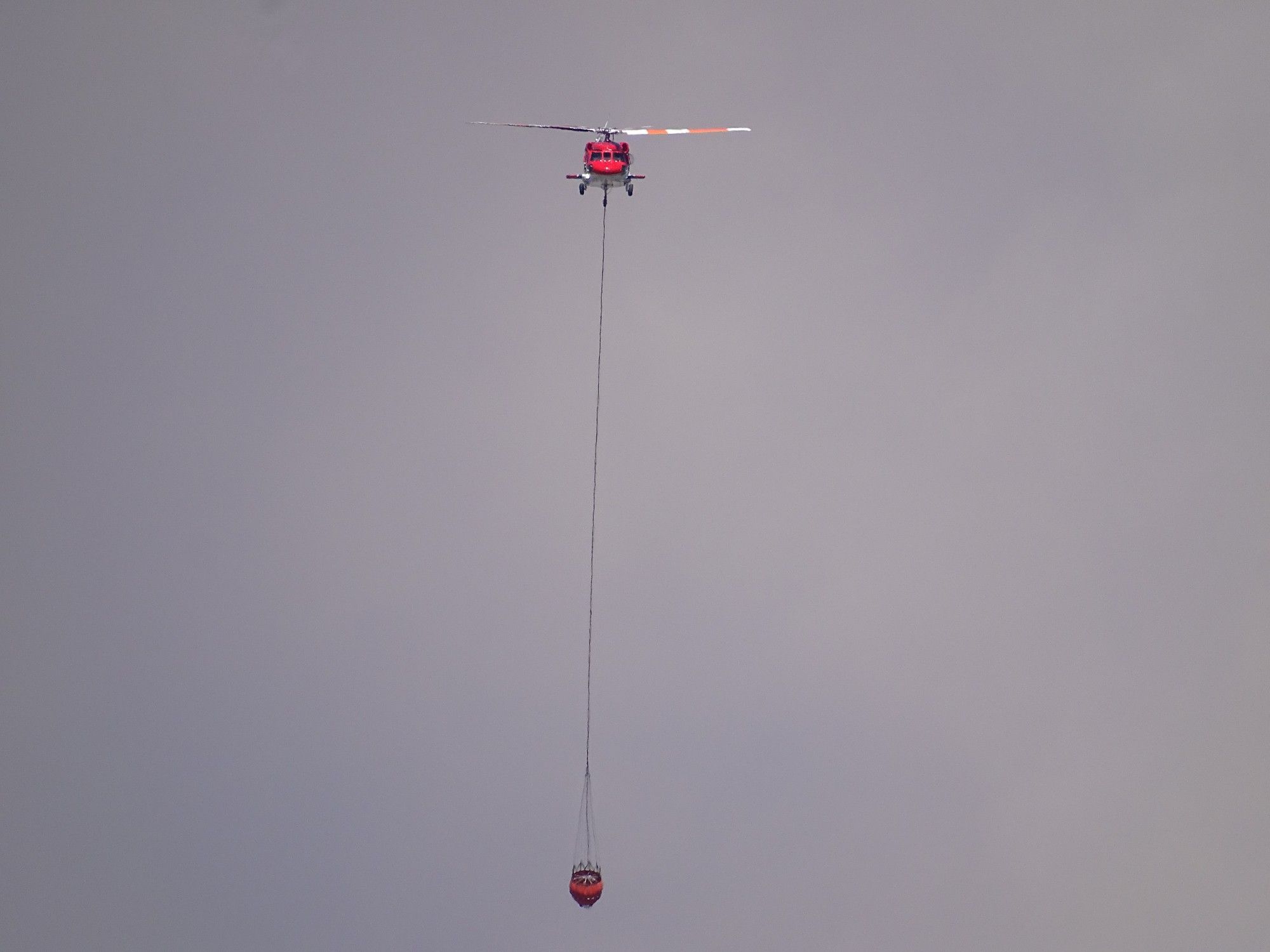 The front view of a red and white Sikorsky UH-60A Black Hawk helicopter as it flies straight down the valley, thick sunlit grey smoke behind it and a large red bucket dangling from a cable below it. The blades are painted an alternating red and white, the nose is red, and the belly is white.