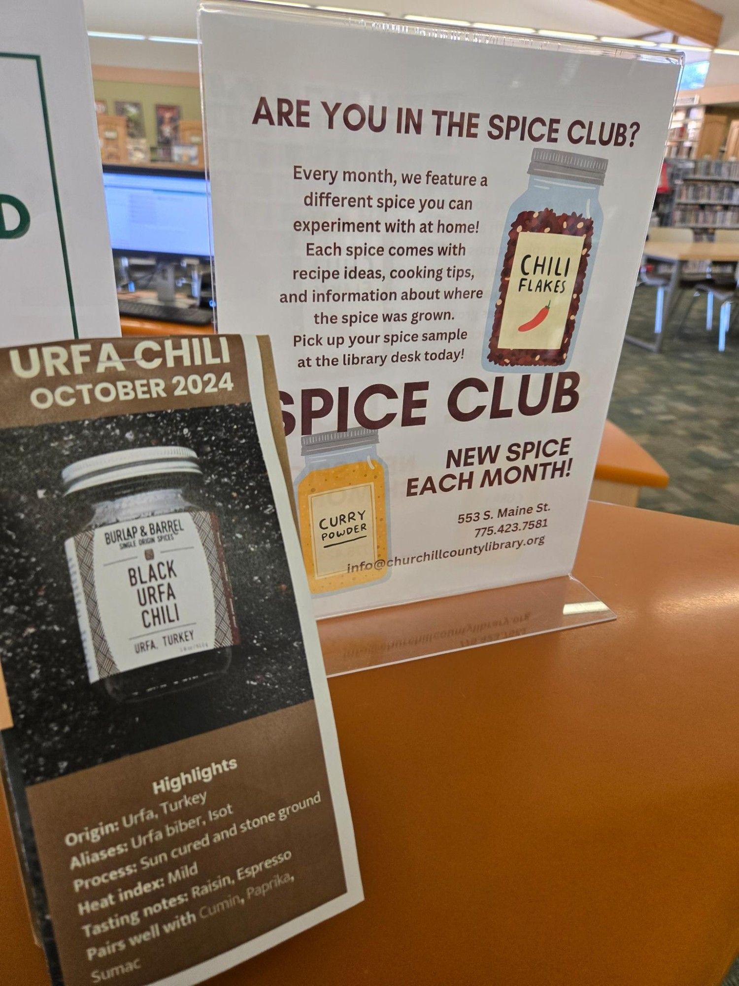 A small brown paper bag with a pamphlet on it that shows a jar of Black Urfa Chili laying on the loose spice. Top of the paper reads "Urfa Chili October 2024" and the bottom shows highlights:
Origin: Urfa, Turkey
Aliases: Urfa biber, Isot
Process: Sun cured and stone ground
Heat index: Mild
Tasting notes: Raisin, Espresso
Pairs well with: Cumin, Paprika, Sumac

A display behind it talks about the club and has clip art of a jar of Chili Flakes and Curry Powder.

Are you in the Spice Club?
Every month, we feature a different spice you can experiment with at home! Each spice comes with recipe ideas, cooking tips, and information about where the spice was grown. Pick up your spice sample at the library desk today!

Spice Club
New Spice Each Month!
553 S. Maine St
775-423-7581
info@churchillcountylibrary.org