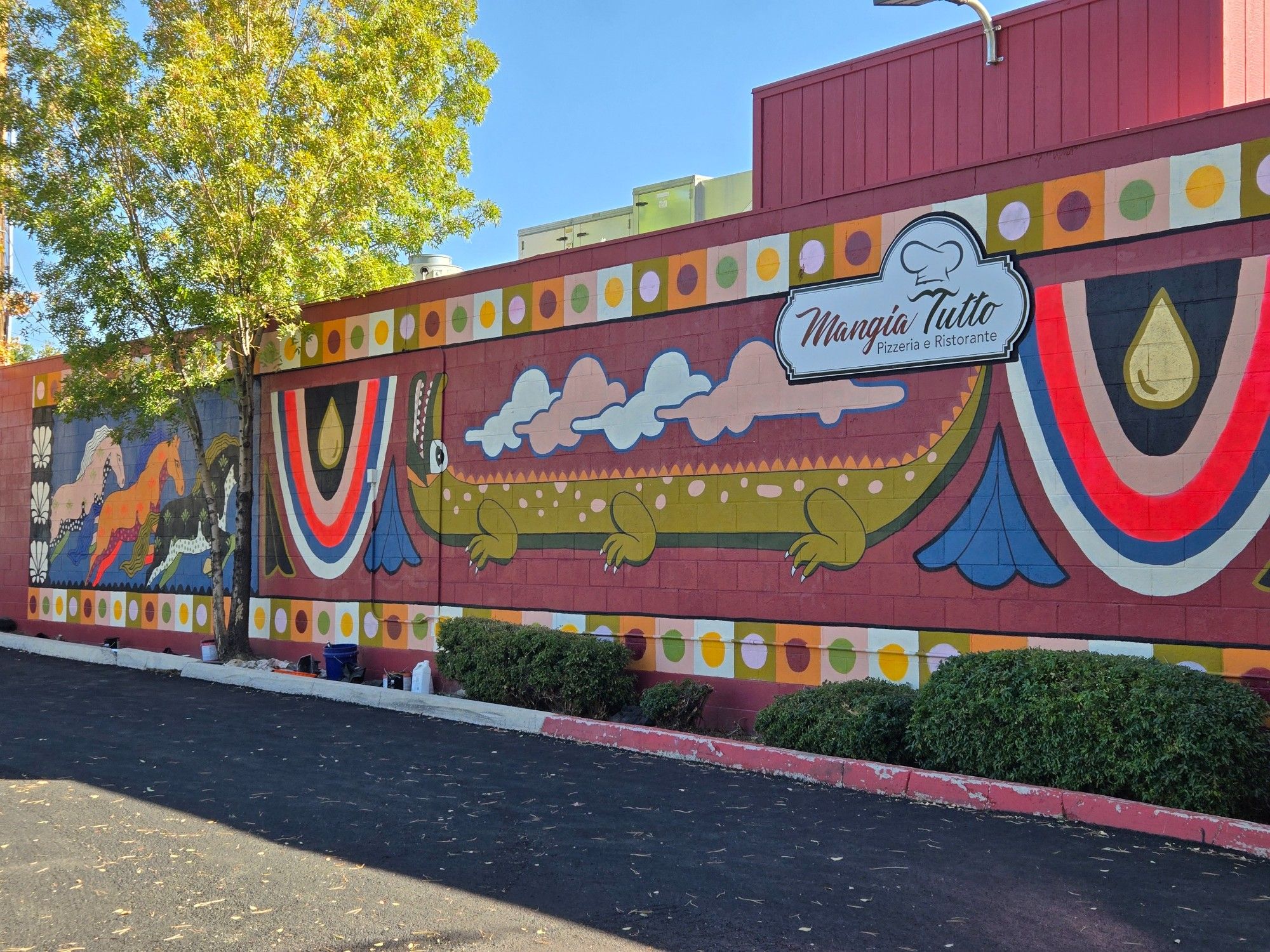 A mural by Brooke Brazil and Co @brookebrazil on the back wall of Mangia Tutto Pizzeria e Ristorante

A long mural resembling the geometric designs of Central America, with three running horses on either end (the right side cut off in the image), the outside one a pale tan, the center one orange, and the inside one black. Each have a plant like design on them and a contrasting color for their shaded bellies. In the center of the mural are two large upside down rainbows, with a large yellow water drop in the middle. The colors are black, tan, orange, blue, and white. Between them is a green stretched six-legged crocodile, with it's head and tail lifted. Pink and blue clouds are above the animal. Around the edge of the image is a row of colored squares with different colored circles in the middle.

The mural is on the west wall and, due to the timing of the visit, is in shadow from the morning sun. The picture is taken at an angle to avoid the sunlight shining on the lens.