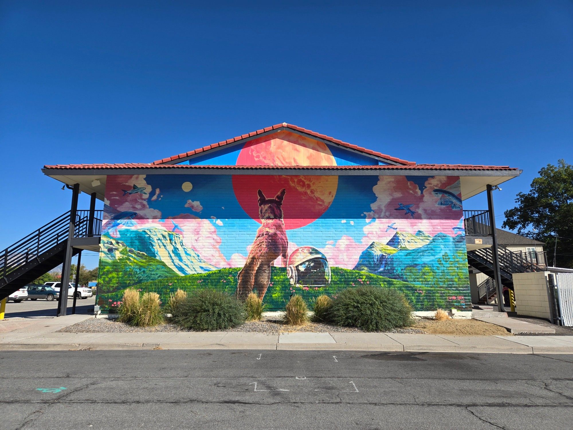 A mural on the side of 323 Flats, by Jabi Corte @jabi_corte

A large red moon hangs over a flowery meadow with large snow capped mountains on either side and fluffy pink clouds in the distance. Standing in the meadow, beside a space helmet set on the ground, is a large brown dog, facing towards the moon. Green trees surround the meadow and blue flying fish swim in the air above the dog.

The artist cleverly used the upper roof to give more size to the moon, the image is taken so the two portions align correctly.