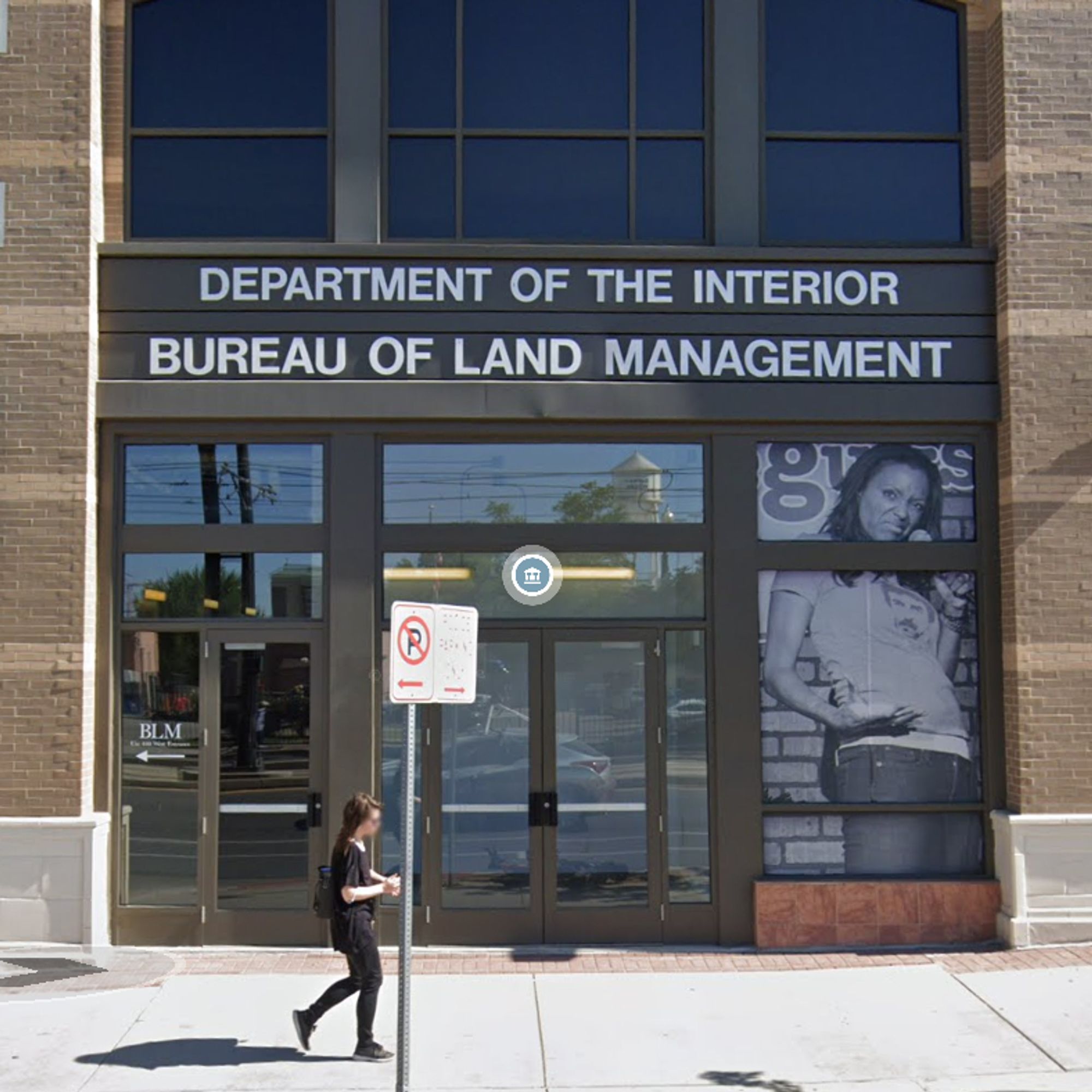 An image from Google Maps Street View shows a woman walking in front of a storefront in downtown Salt Lake City. The sign over the entrance states, in all-caps, Department of the Interior Bureau of Land Management. Under the sign is a large, monochromatic window graphic depicting the comedian Aisha Tyler performing at the Wiseguys Comedy Club.