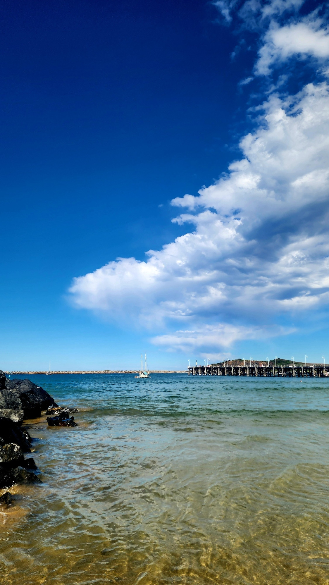 A beach shot taken from the shallows looking out to sea. Dark rocks on the left. The sky is vibrant blue with bright white clouds. A sailboat appears tiny in the distance. 