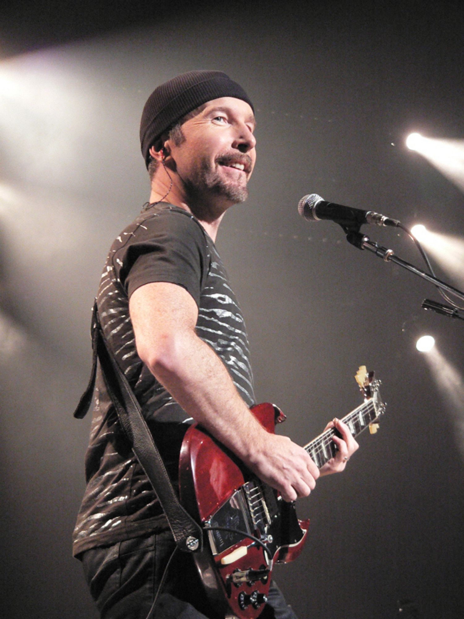 A profile photo of The Edge, from the band U2, holding a red electric guitar and smiling toward the crowd. The shot is lit by multiple white spotlights.