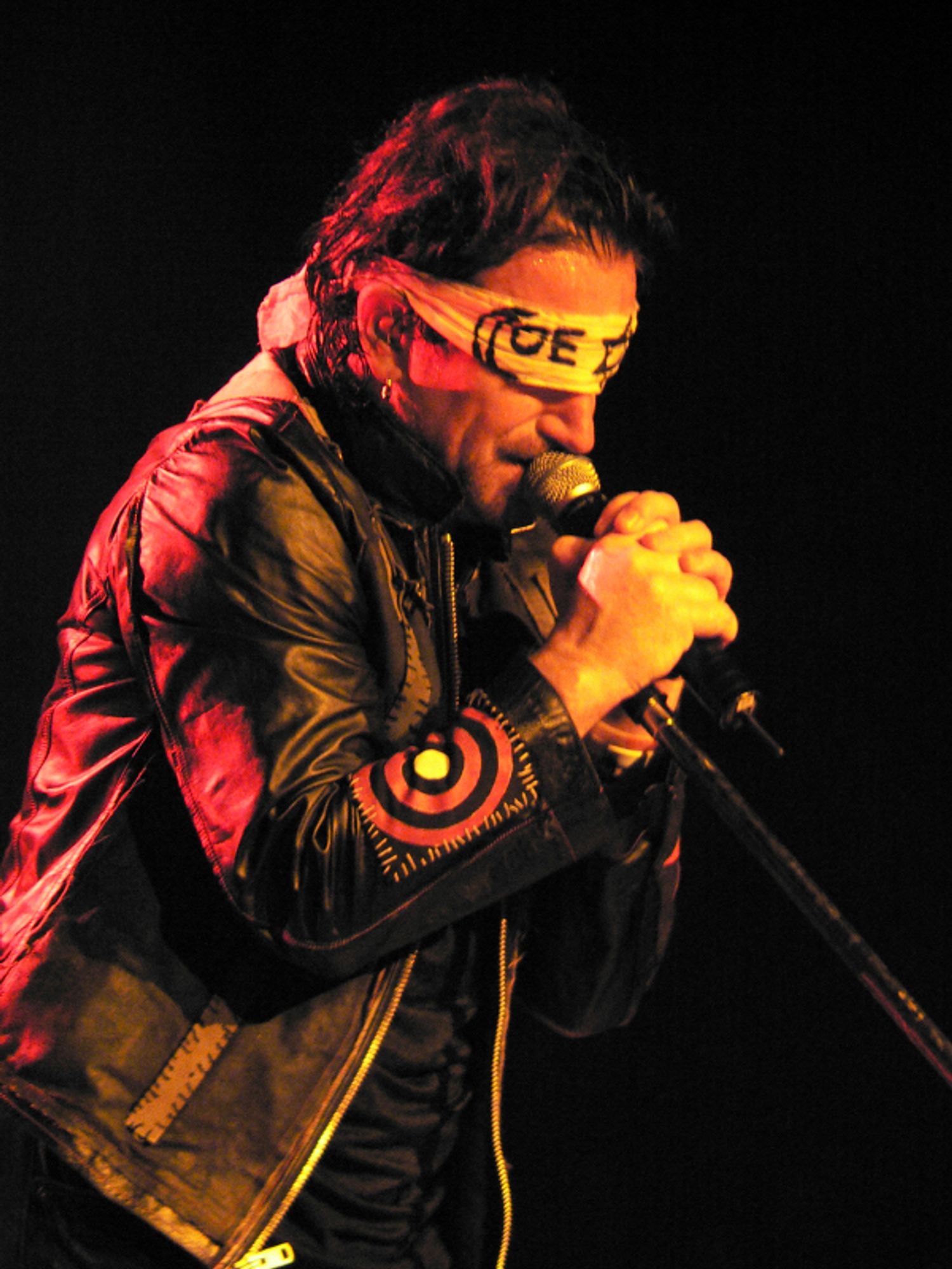 Bono, of the band U2, holding a microphone in two hands and wearing a leather jacket and a “Coexist” bandana as a blindfold. The singer is lit in red and orange stage lighting.