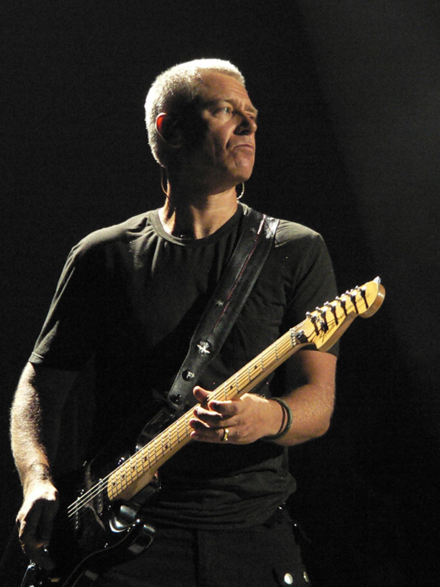 Adam Clayton, of the band U2, wears all black and strums his bass guitar in a dark photo. He is lit by two(?) warm spotlights and he looks out into the crowd.