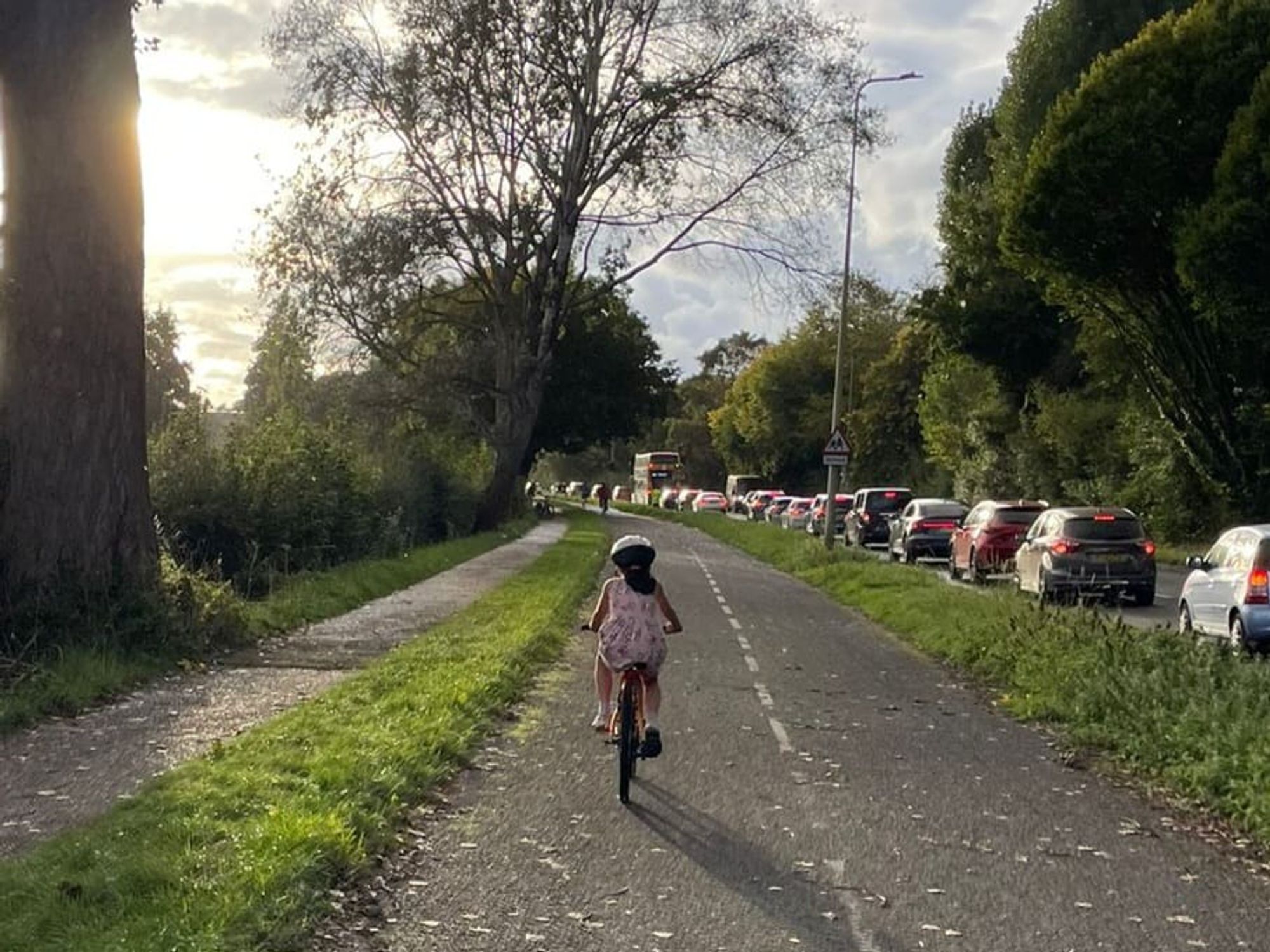 A little girl on a bicycle is seen on an open bicycle path, with her back to the camera, next to a traffic jam of stationary cars. Sunlight dapples the path.