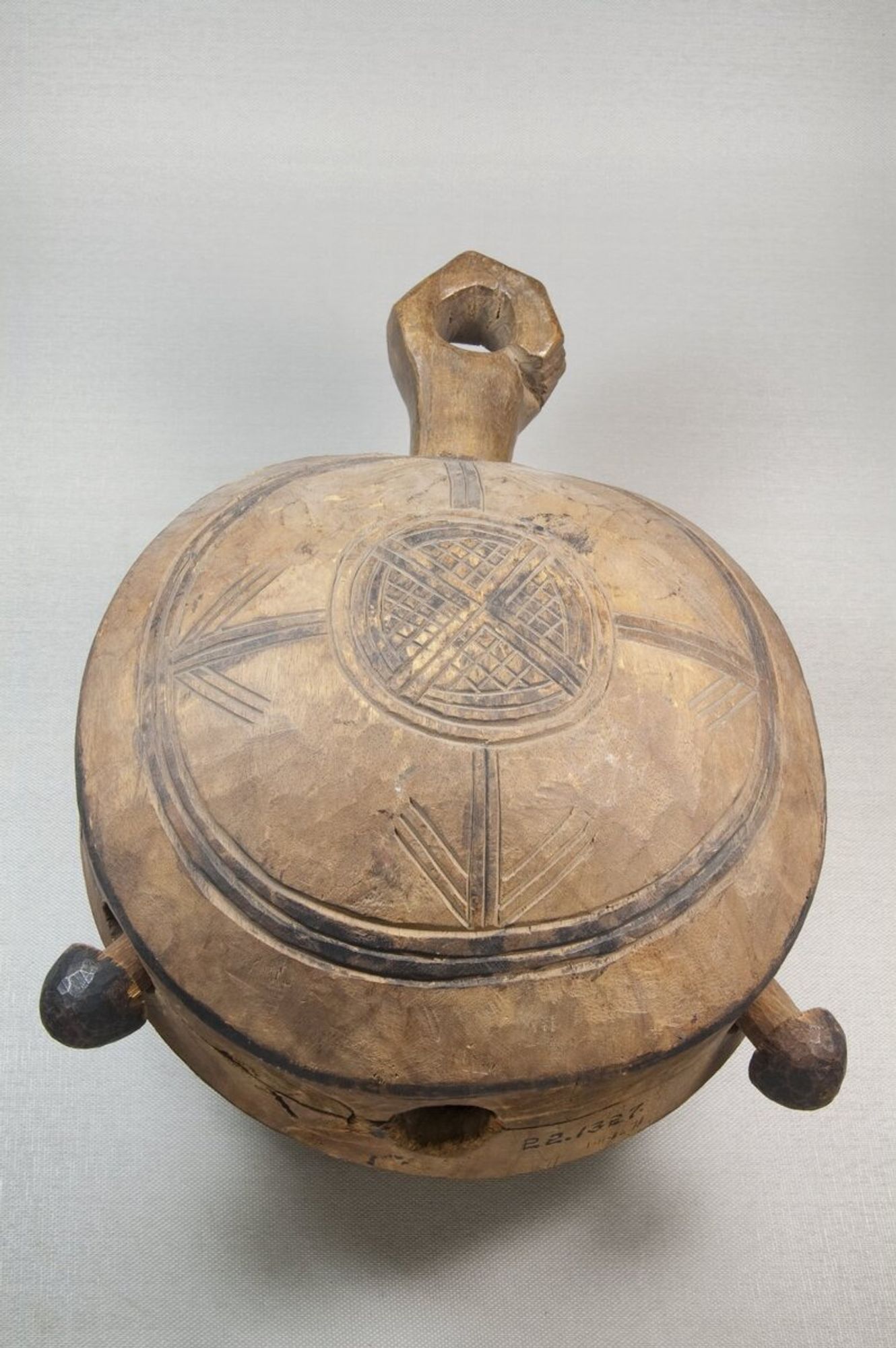 Circular wooden bell carved on both sides with handle in form of a human hand. One side decorated with circle inside of which are incised squares and straight lines; on other side there is a circle within a circle with four arrows radiating from inner to outside circle.  There are two projectons as clappers.  Brown  with black decoration.  CONDITION: Very worn, one clapper mising; hardened black lacquer-like substance on one side; nicks and gouges throughout.