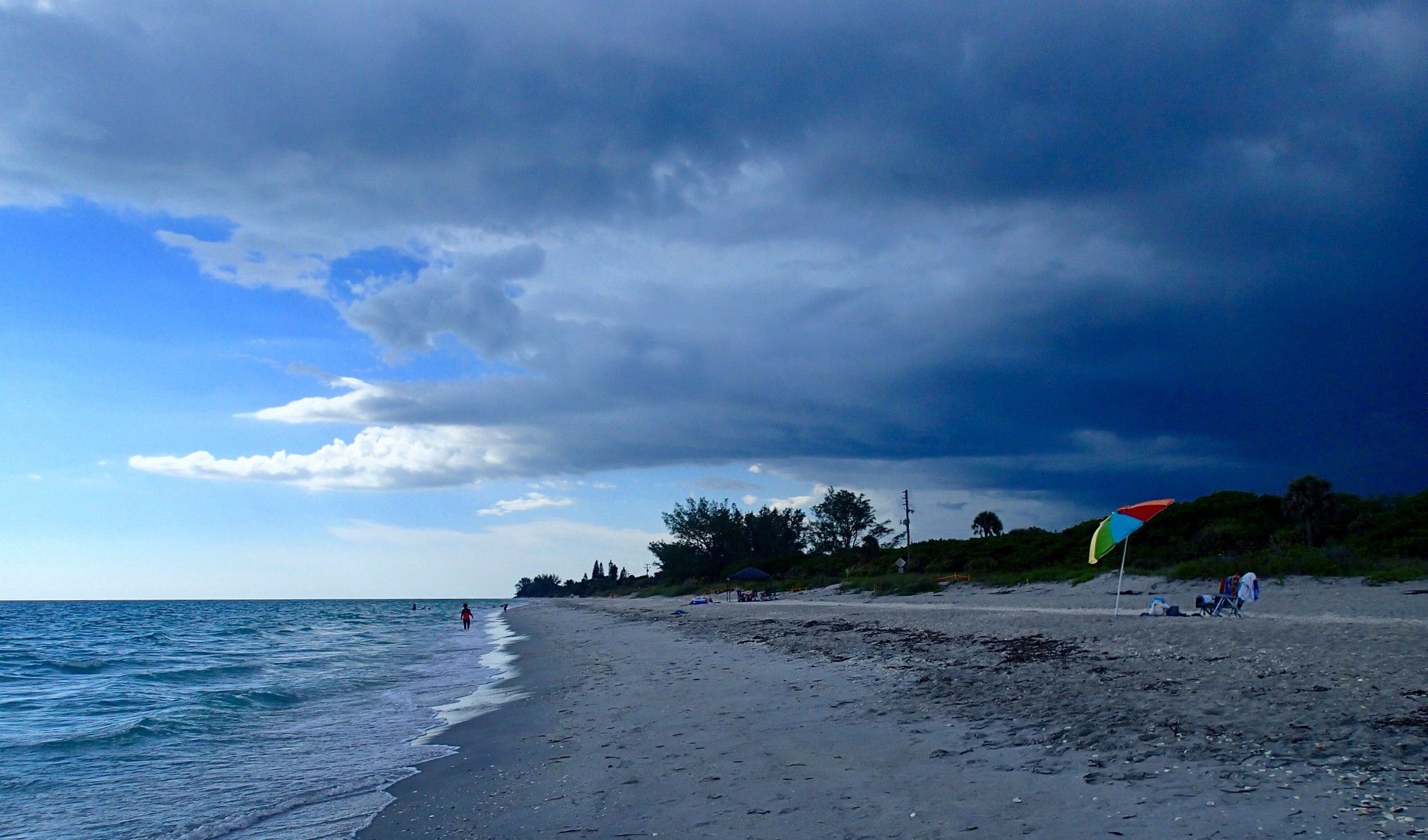 Photo taken at Blind Pass Beach of a sky full of dark blue clouds on the right. Lighter blue and white clouds on the left. A person wades in the calm Gulf water and other people sit under a rainbow umbrella on the beach, behind the wrack line. Trees and shrubs in the background.