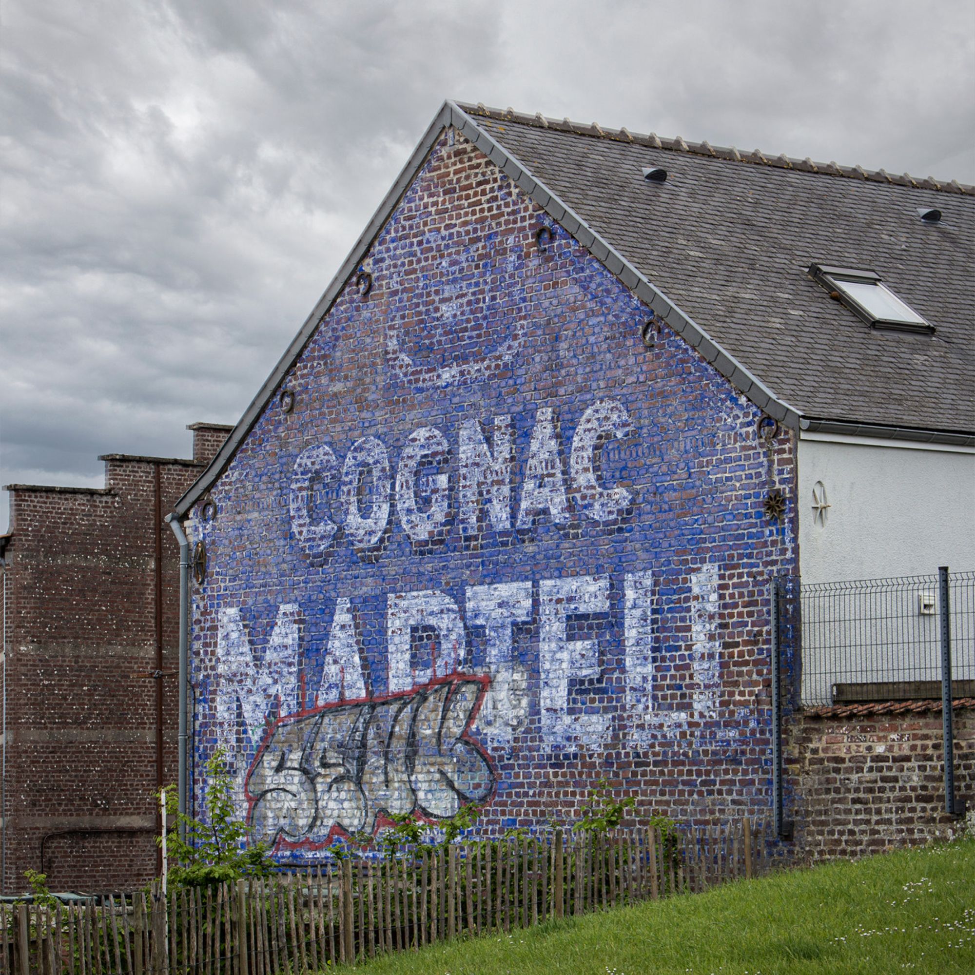 Mur peint en bleu sur le flanc d'une maison : Texte « Cognac Martel », surchargé d'un graffiti « Seuk »