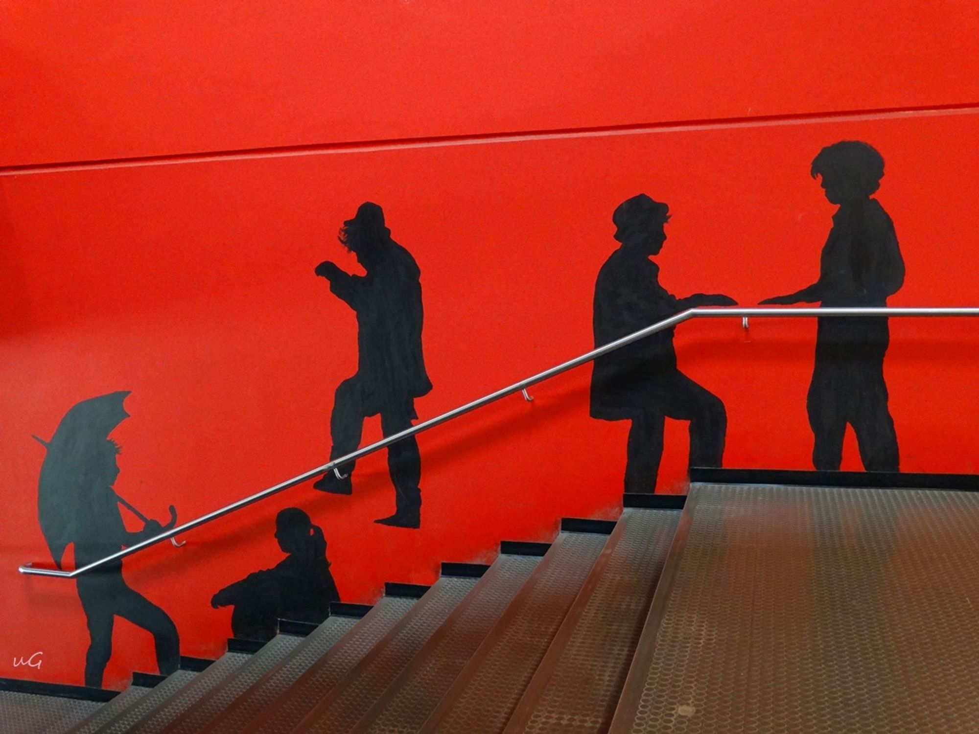 Eine Treppe von der Seite fotografiert, ihre Stufen sind sehr lang. Die begrenzende Wand ist rot angestrichen, darauf sind schwarze Silhouetten im Profil gemalt.
Da es sich um eine Schule handelt, sind die Figuren im Teenageralter.
Von unten nach oben: ein Junge mit einem Regenschirm hat den Fuß auf die erste Stufe gesetzt, ein Mädchen mit Pferdeschwanz hockt auf der vierten Stufe, sie hat die Füße auf der dritten abgestellt, einer mit Hut scheint die Treppe herunter zu hüpfen, er schwebt gerade in der Luft. Auf er obersten Stufe geht einer mit Hut und Mantel und streckt dem ihm entgegen kommenden Jungen, der noch auf der Plattform vor den Stufen steht, die Hand entgegen, so, wie der mit Hut es auch macht.