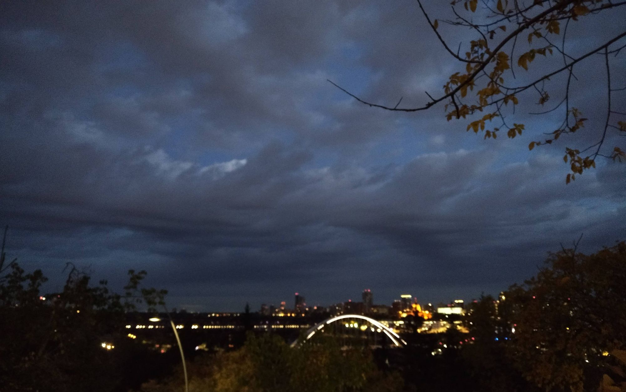 Under a dark twilight cloudy sky, the western edge of downtown is visible in lights through the trees of the river valley.

A long horizontal line of white headlights on the High Level Bridge crosses the river valley from plateau to plateau. A bright white arc of the Walterdale suspension bridge crosses the river in the valley, and the orange glow of an obscured Legislature building glows behind and above it.