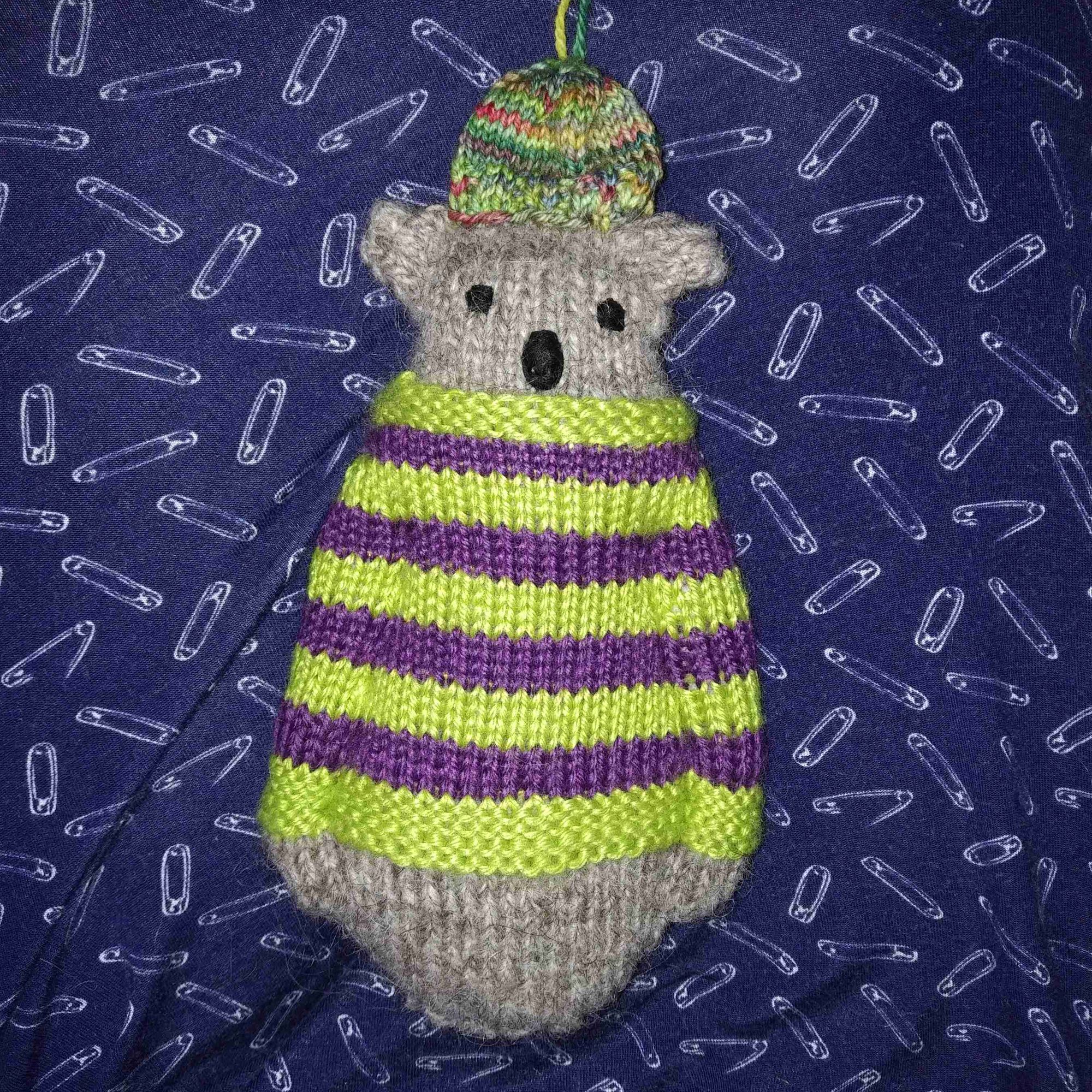 A knit bear tree ornament wearing a purple and green striped sweater and a green and red variegated toque with a hanging string on it.
The bear is grey and pear-shaped with a rounded bottom (and no legs) with its arms denoted by stitched-in divots. Its eyes are uneven and its nose is oblong like a koala's.