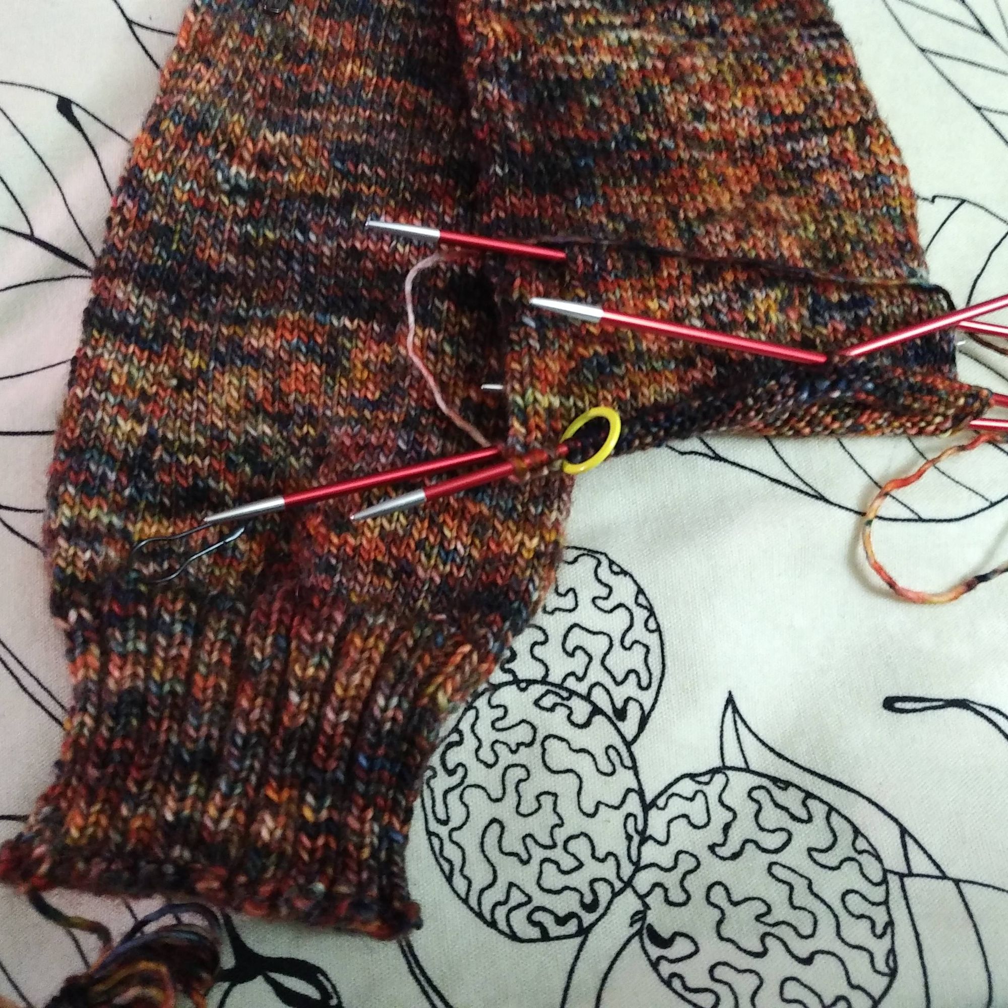 A pair of knit socks in the process of being knit.
One sock, on the left is done. One sock, on the right, is in process, with knitting needles still in it.
The socks are in a firey orange and red colour design with flecks of black/navy and white in it.