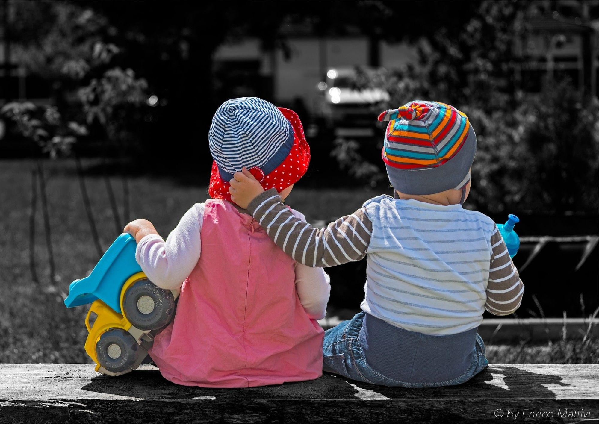 Zwei Kleinkinder von hinten sitzend auf einer Bank als Colorkey. Der Junge legt ganz schüchtern seinen Arm um das Mädchen., welches sein Spielzeug LKW in der Hand hält.