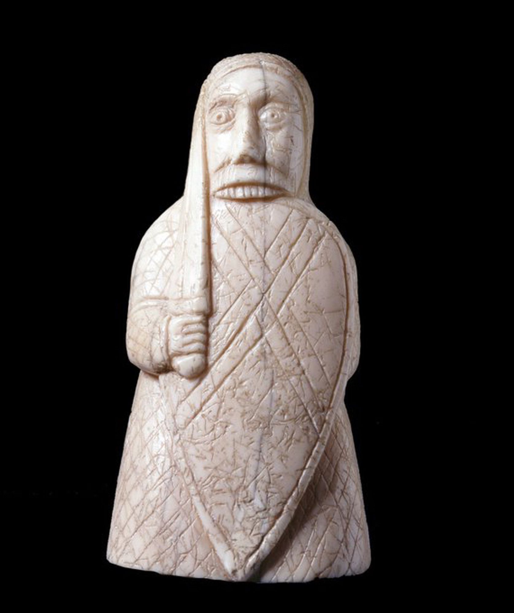 One of the Lewis Chessmen. Carved from walrus ivory, probably in the 12th century. This chesspiece depicts an armoured warrior – quilted armour or perhaps chain-mail is suggested by diagonal cross-hatched lines inscribed into the long coat, which also forms a hood over the character's head. He holds a full-length kite-shaped shield in front of him, bearing an X-shaped cross made of four interwoven lines. Holding a sword in his right hand, clutched close against his body, the warrior's outsized face stares over the top of the shield. His top set of teeth are visible, chewing at the shield-top in Berserker battle-frenzy.