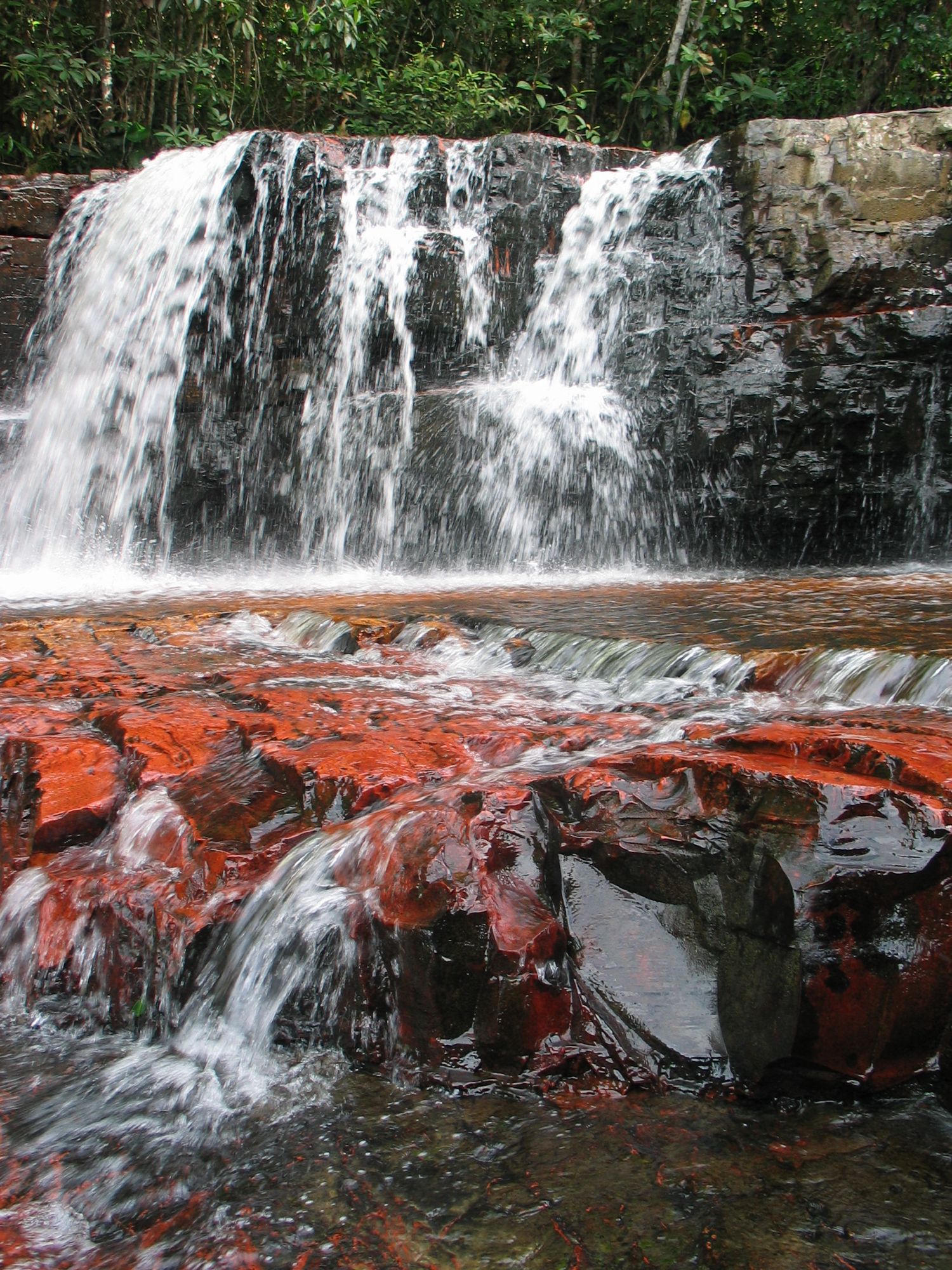 Ein Wasserfall stürzt über schwarzen Stein. Im Vordergrund ist eine Stufe aus rötlichem Stein, über die das Wasser flach läuft.