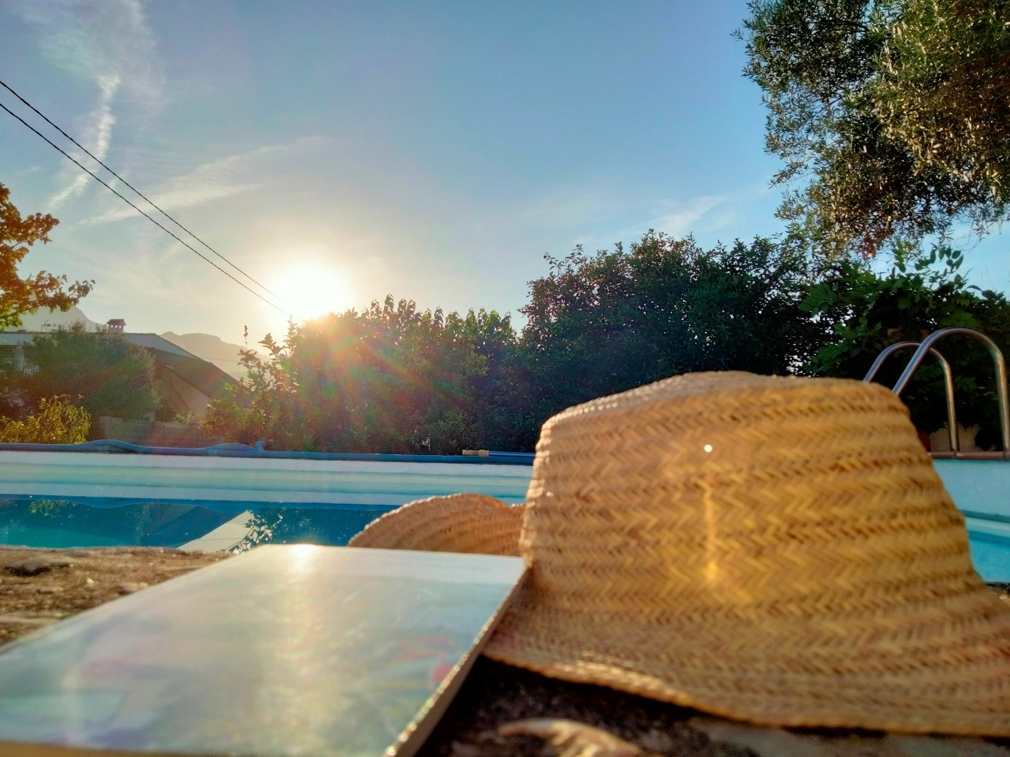 Fotografia de un sombrero y un libro en primer plano. Detrás de ellos una piscina y más al fondo unos árboles. Detrás de los árboles se asoma el sol, que alumbra directamente hacia el frente