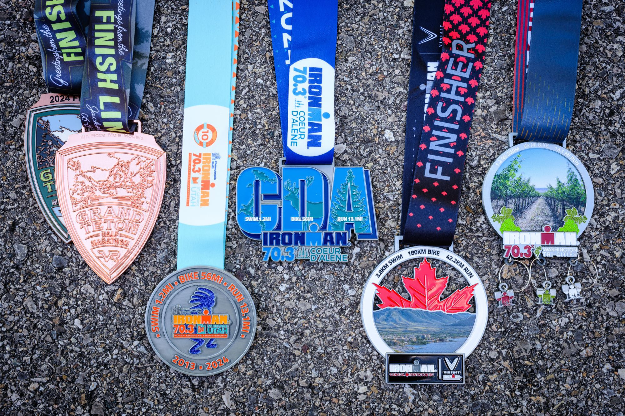 Six medals from various races laid out on pavement. From left to right, the first medal is a finisher's medal for the Grand Teton Half Marathon; the second medal, on top of the first one, is a bronze "4th Place Male Masters" medal for the same race; the third medal is from Ironman 70.3 St. George; the fourth medal is from Ironman 70.3 Coeur d'Alene; the fifth medal is from Ironman Canada and features a red maple leaf and a mountainous landscape; the sixth medal is from Ironman 70.3 Washington Tri-Cities, featuring a landscape with vines, and three wine glass charms with the M-dot logo at the bottom.