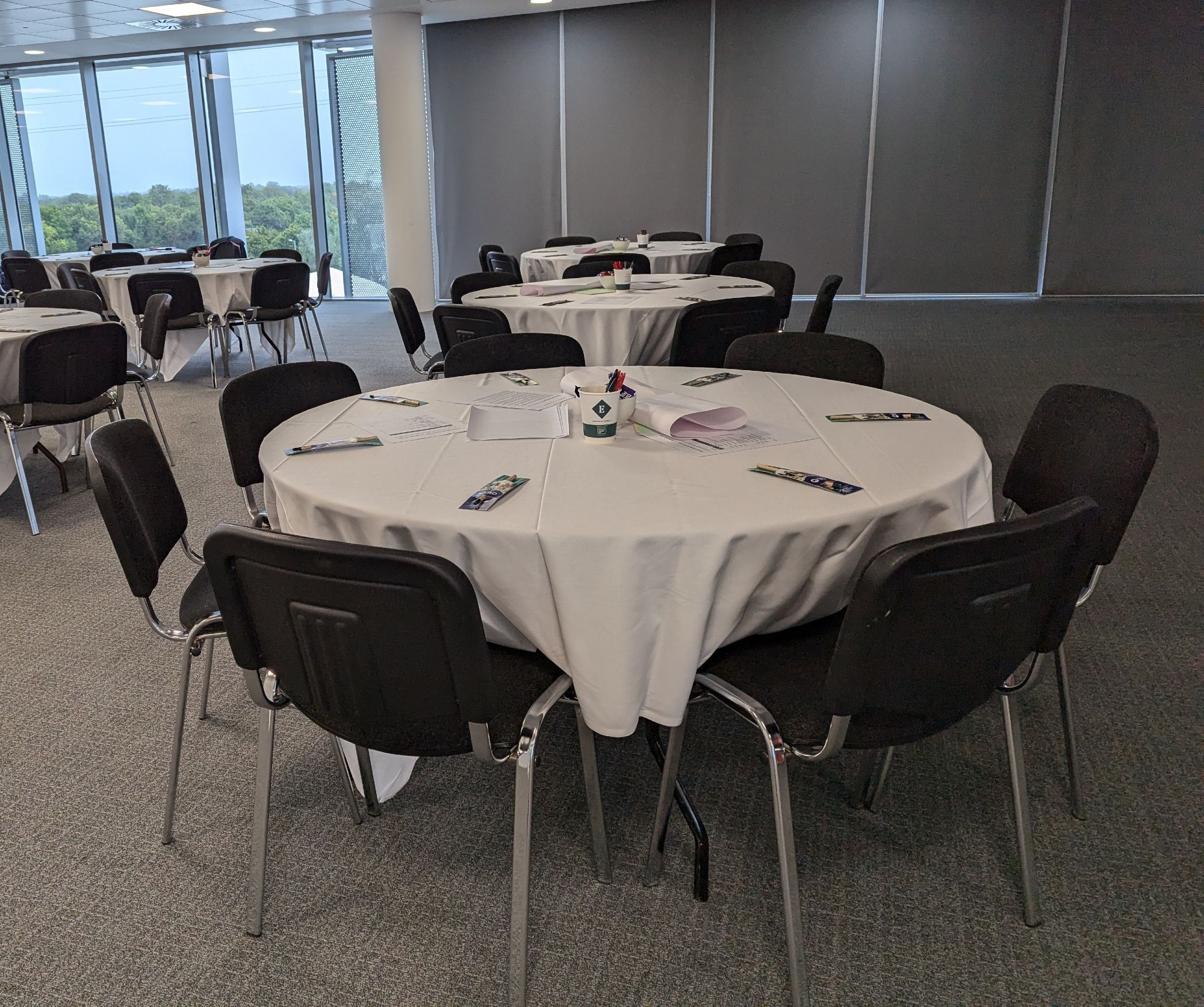 A photo of a set of round tables with pens and paper, each surrounded by seven chairs and pens.